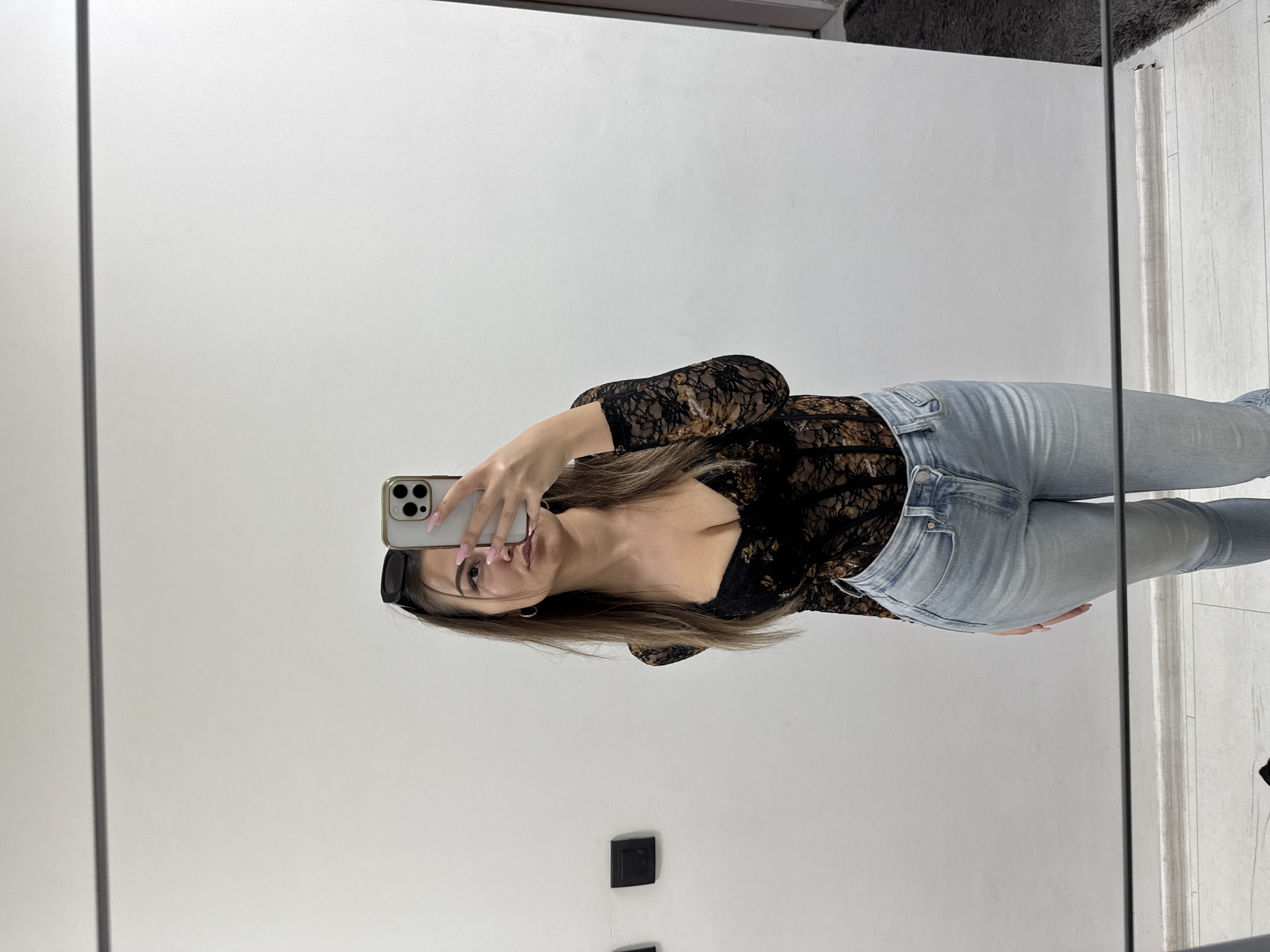 This mirror selfie, taken in Sofia, Bulgaria, captures a young woman standing indoors. She has long, straight dark brown hair, parted in the middle, and wears black sunglasses pushed up onto her head. Her expression is neutral as she looks directly into her smartphone, which she holds up with her right hand to take the picture. Her manicured nails are painted a light pink.
She is dressed in a fitted, black long-sleeved sheer lace top featuring a subtle floral or leafy pattern, with a low V-neckline. This is paired with light-wash blue skinny jeans.
The background, as reflected in the mirror, consists primarily of a plain, bright white wall. Towards the bottom right, a segment of a light-colored wooden or laminate floor is visible. Two dark square objects, possibly electrical outlets or decorative elements, are mounted on the lower part of the white wall. A dark, possibly furry, object is partially reflected in the top right corner, while a thin dark vertical line on the left suggests the edge of the mirror frame or a structural element of the room. The lighting appears bright and even, indicating it is likely daytime. There is no visible text in the image.