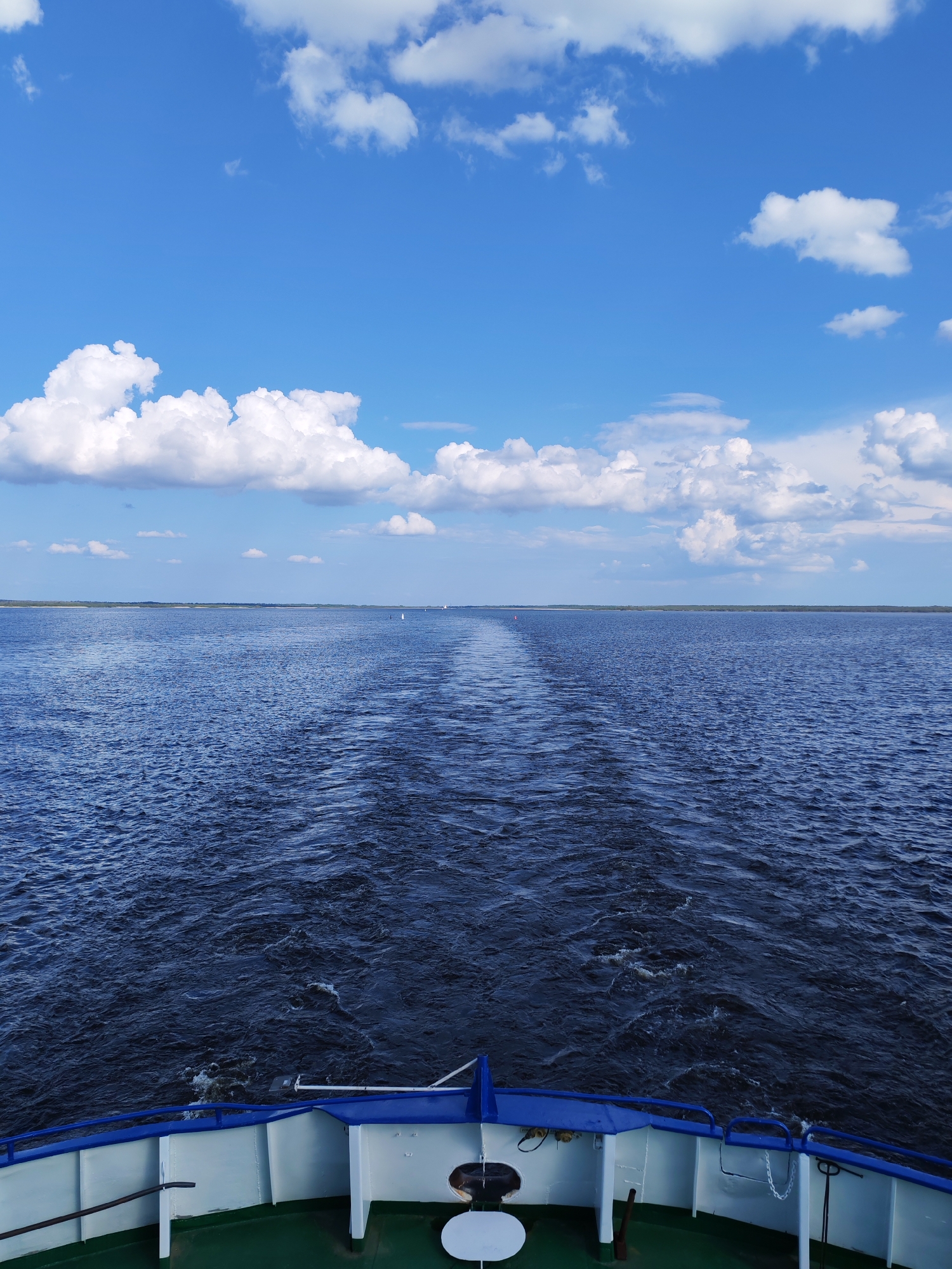 The image depicts a view from the stern of a vessel traveling on water, likely a river or a large lake. The wake of the vessel is visible, stretching towards the horizon. Beyond the water, a distant shoreline is faintly discernible. The sky is predominantly blue with scattered white clouds. In the foreground, parts of the vessel's railing and deck are visible.
There is no verifiable information within the image to confirm that the location is Veregonets, Russia.