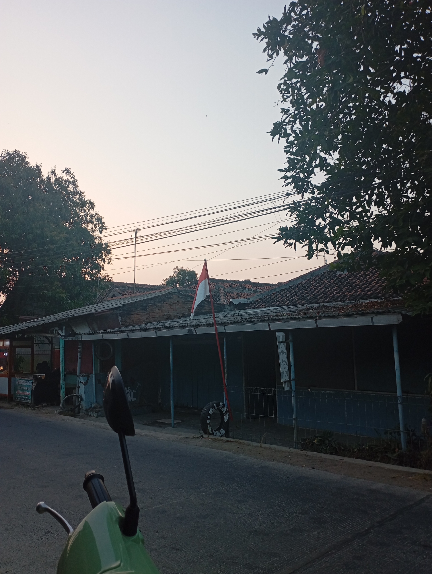 This is a street-level, eye-level shot taken from a motorcycle, looking towards a row of buildings. The scene is set in Kecamatan Haurgeulis, Indonesia, possibly in the early morning or late afternoon given the soft, warm light and the clear sky with subtle pastel hues. In the foreground, the green handlebars and a black rearview mirror of the motorcycle are visible. The road in front is asphalt and appears to be in good condition.
The middle ground features a long, single-story building with a tiled roof that appears aged. The building has a covered porch area supported by blue pillars. A red and white flag, likely the Indonesian flag, is prominently displayed on a pole in front of the building. Next to the flag, a used tire stands upright, and to its left, a section of a metal fence is visible. The building's facade is partially obscured by shadows and appears to house some sort of establishment with signs that are difficult to read clearly, though one sign on the left seems to display some text in Indonesian.
The background is dominated by lush green trees on the left and right, framing the view of the sky. Power lines stretch horizontally across the upper portion of the image, indicating the presence of infrastructure. The overall atmosphere is calm and tranquil, suggesting a quiet street scene in a residential or commercial area of the town. There are no clearly discernible people in the image, though the shadows suggest some activity within the building.