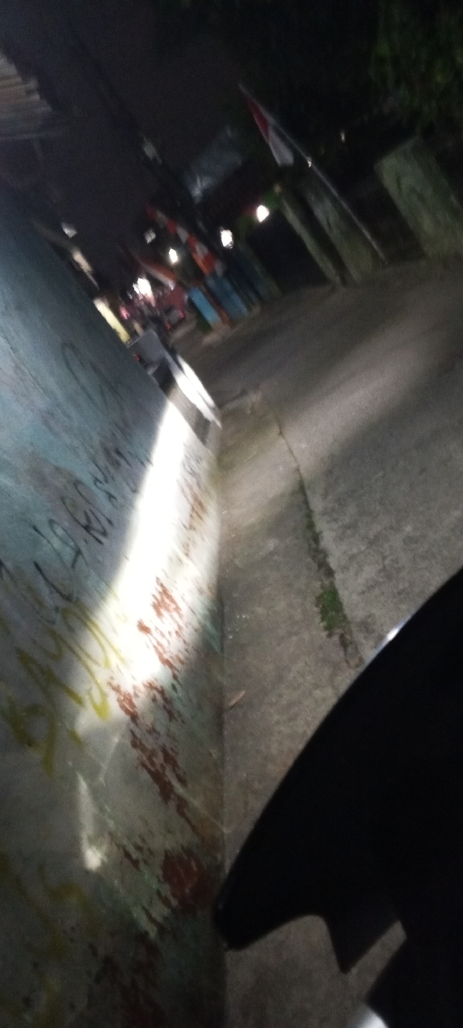 The image, taken at night in Depok, Indonesia, shows a narrow street or alleyway illuminated by the headlights of a vehicle, presumably a motorcycle given the visible portion of a black fender in the foreground. The left side of the frame is dominated by a graffiti-covered wall, with distinct patches of red and yellow spray paint. The wall appears to be made of concrete or plaster. To the right of the wall is a paved surface, possibly a sidewalk or road, which is also illuminated by the headlights. In the background, several structures are visible, including what appears to be a roof with some decorative flags or banners. There are also indications of other vehicles and possibly people further down the alley, but they are indistinct due to the low light conditions. The overall impression is of a dimly lit urban environment, characteristic of residential or commercial side streets in Indonesia at night.