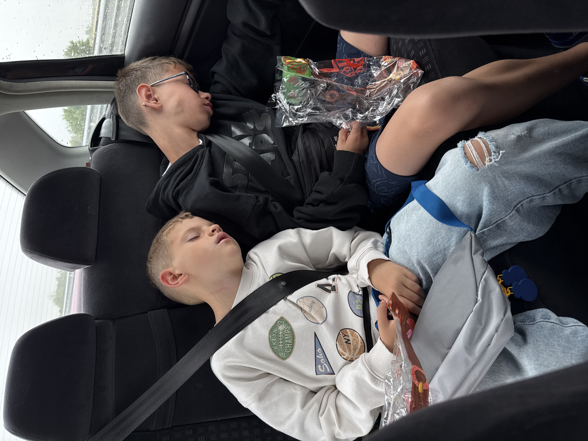 Two young males are asleep in the back seat of a vehicle. The male on the left is wearing a white sweatshirt with various patches and light-wash ripped jeans. He is resting his head on a gray fabric object. The male on the right is wearing a black hoodie and dark patterned shorts. He has glasses on and is holding a clear plastic bag containing what appears to be candy. Both males are wearing seatbelts. The background shows the interior of a car, with the rear seats and headrests visible. Through the window, a blurry, overcast sky and some greenery can be seen, suggesting it is daytime and possibly raining. The scene is consistent with individuals sleeping during a car journey. The image does not contain any specific visual cues that would place it in Malo Konare, Bulgaria.