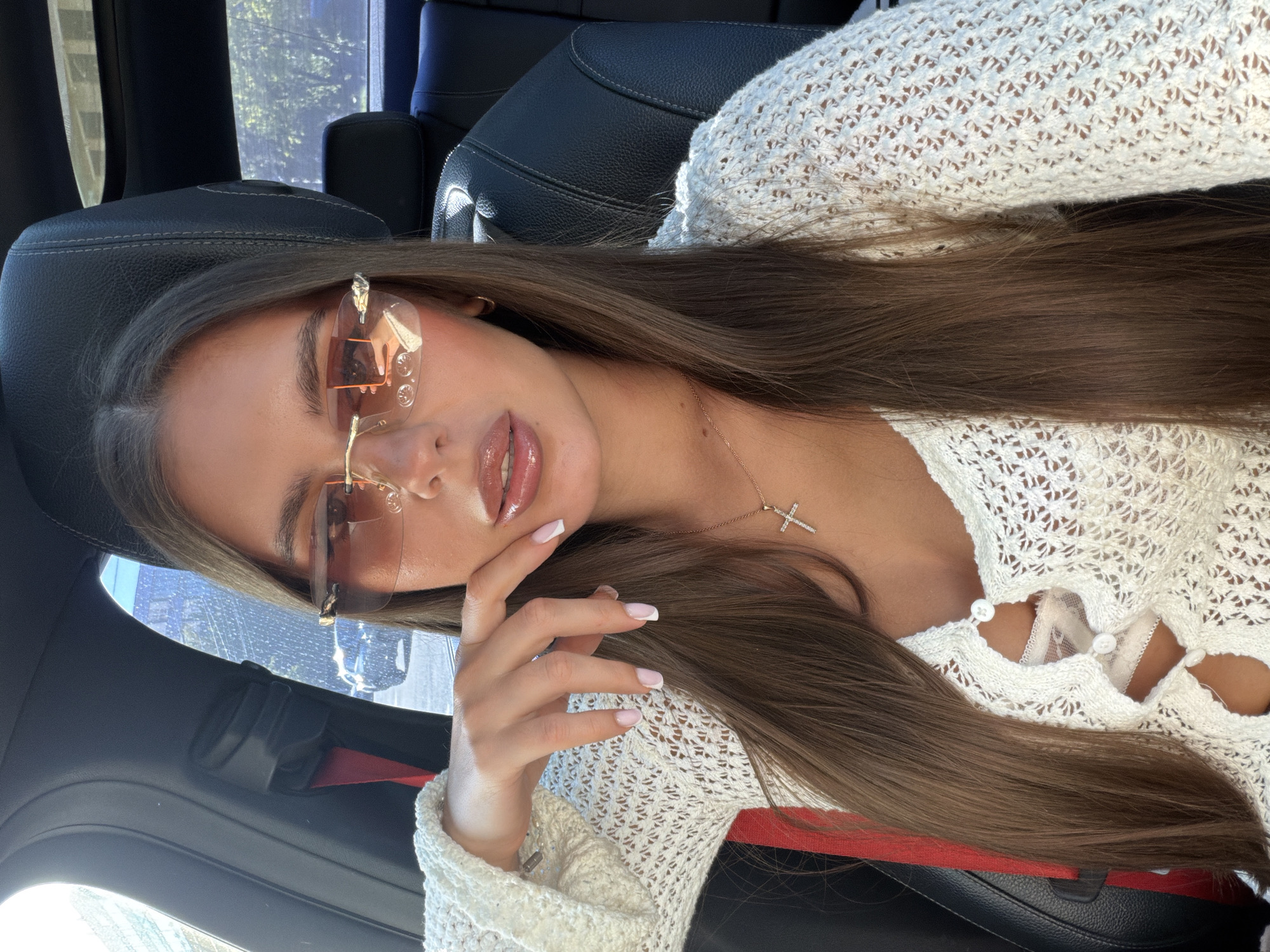 A young woman is depicted in a close-up, slightly tilted portrait shot, seated in the passenger side of a car. She has long, straight brown hair, parted down the middle, cascading over her shoulders and part of her chest. She wears rimless sunglasses with orange-tinted lenses, reflecting elements of the car's interior. Her lips are full and glossy, and her left hand, adorned with French-tipped nails, is gently touching her chin.
She is dressed in a white, open-knit sweater or cardigan with visible buttons down the front, revealing a white lace bra or bralette underneath. A delicate gold-toned chain with a small cross pendant hangs around her neck. The car's interior is primarily black, with leather seats visible behind her and a glimpse of a red seatbelt component. Through the car windows, some green foliage and bright outdoor lighting can be discerned, indicating it is daytime. The scene suggests a casual moment, possibly a selfie or posed photograph, within a vehicle in Krasnoyarsk, Russia. There is no visible text in the image.