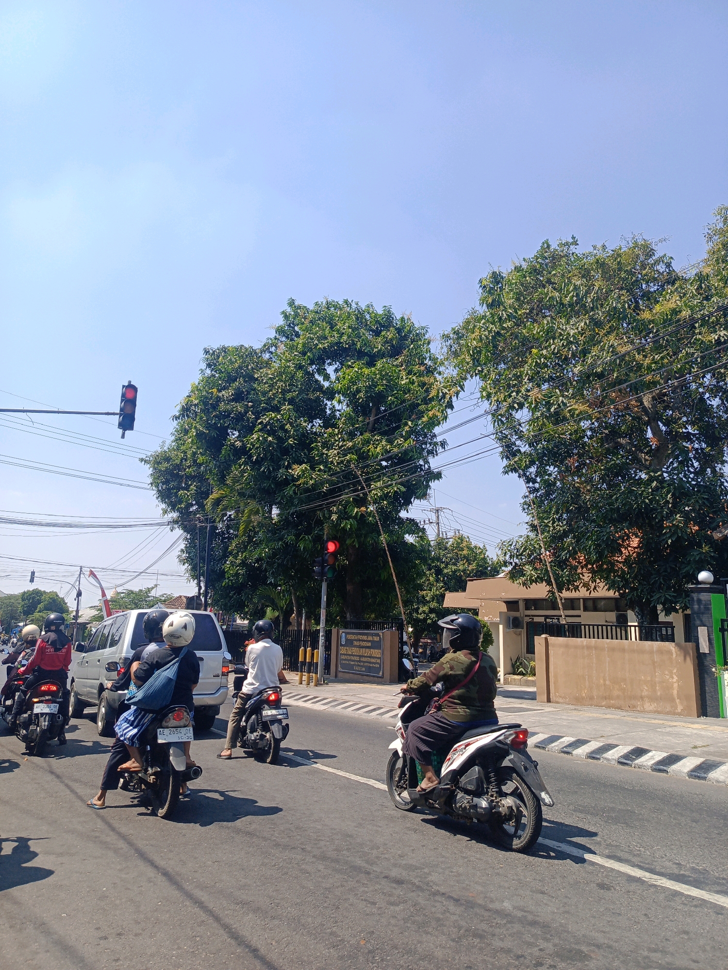 The image depicts a street scene in Sukowinangun, Indonesia, under a clear blue sky on a sunny day. The foreground shows a paved road with several motorcycles and a silver van. On the road, there are multiple people riding motorcycles. One person in the foreground is riding a white motorcycle, wearing a dark green jacket, black pants, and a black helmet, with a red strap across their chest. Behind them, a person in a blue bag and striped pants is riding a white motorcycle with license plate AE 2654 DE. To the left, two people are on a red motorcycle, and a silver van is stopped. Further to the left, another motorcycle with a rider in a red jacket is visible. In the background, there are large green trees and a building with a light brown facade, fenced with black metal bars. A traffic light with a red signal is visible on a pole extending over the road, and another traffic light with a red signal is attached to a pole near the building. The road has white lane markings and a sidewalk with black and white striped crosswalk markings. The overall scene suggests a typical urban traffic environment in Indonesia.