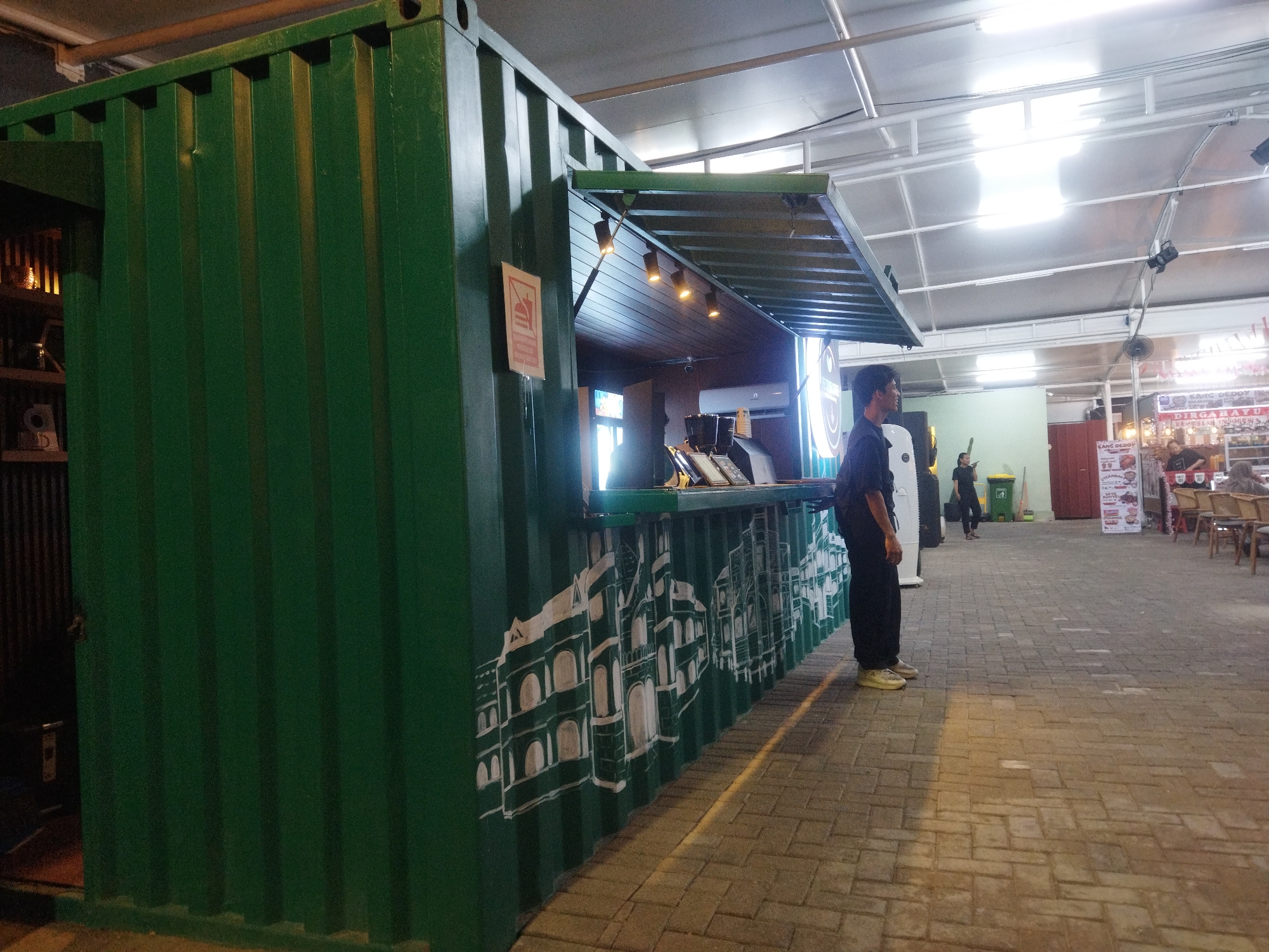 A green shipping container repurposed as a food stall is depicted. The exterior of the container features a mural of white architectural drawings, possibly of European buildings. A man in black clothing and yellow shoes stands to the right of the stall, facing left. The stall has an open serving window with lights above, and a counter inside. The ground is paved with interlocking bricks. In the background, other stalls, tables, chairs, and people can be seen, suggesting a market or outdoor dining area. The scene appears to be located in a covered outdoor space, possibly a food court or night market. The lighting suggests it is either evening or a dimly lit indoor space. The context of Semarang, Indonesia, suggests this is likely an urban market or eatery.
