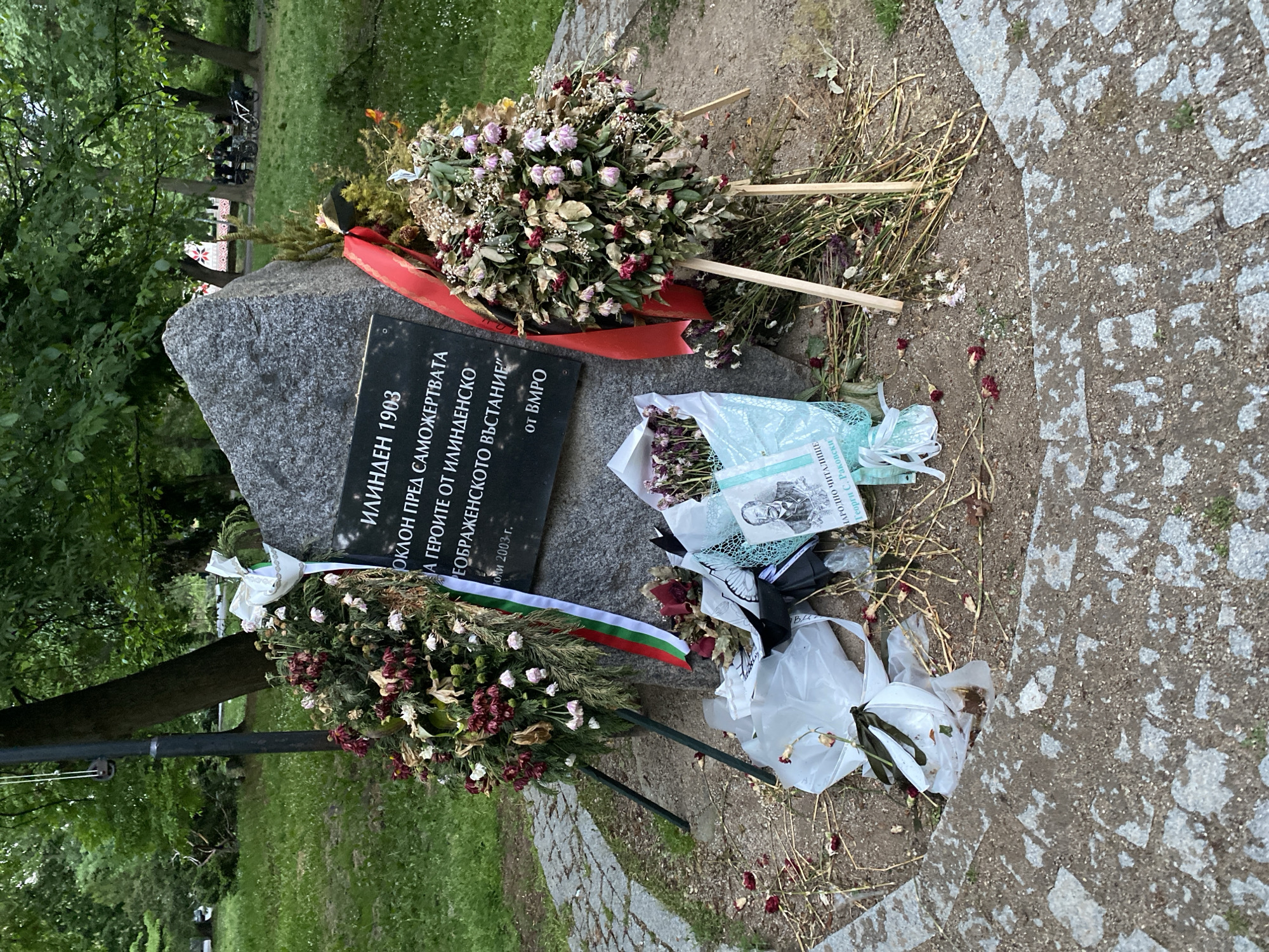 The image shows a memorial stone dedicated to the Ilinden-Preobrazhenie Uprising of 1903, located in the city of Sofia, Bulgaria. The gray stone is inscribed in Bulgarian with the year "1903", "ИЛИНДЕН 1903" and text regarding the self-sacrifice of the heroes. Several wreaths of flowers, both fresh and decaying, adorn the stone, along with a Bulgarian flag ribbon. Discarded plastic wrapping and other debris litter the area around the memorial, contrasting with the solemnity of the site. The stone is positioned on the edge of a paved area, with grass and trees visible in the background, suggesting a park setting.