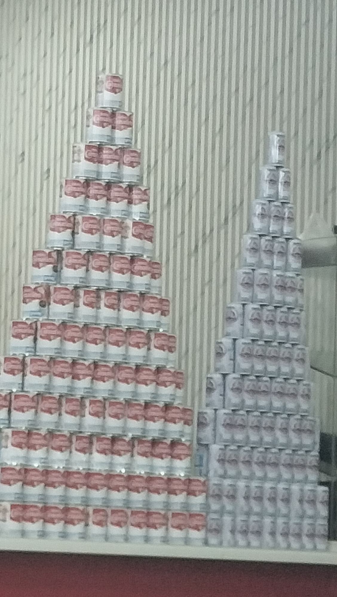Two pyramids of canned goods are displayed against a corrugated metal wall. The larger pyramid on the left is composed of cans with red labels and white tops, stacked in ascending layers. The smaller pyramid on the right is made of cans with white labels. Both structures are arranged on a white surface, with a dark red surface visible at the bottom. The image is blurry, suggesting it was taken from a distance or with a low-resolution camera. There are no people or visible actions depicted in the scene. The location in Sigli, Indonesia is not specified.
