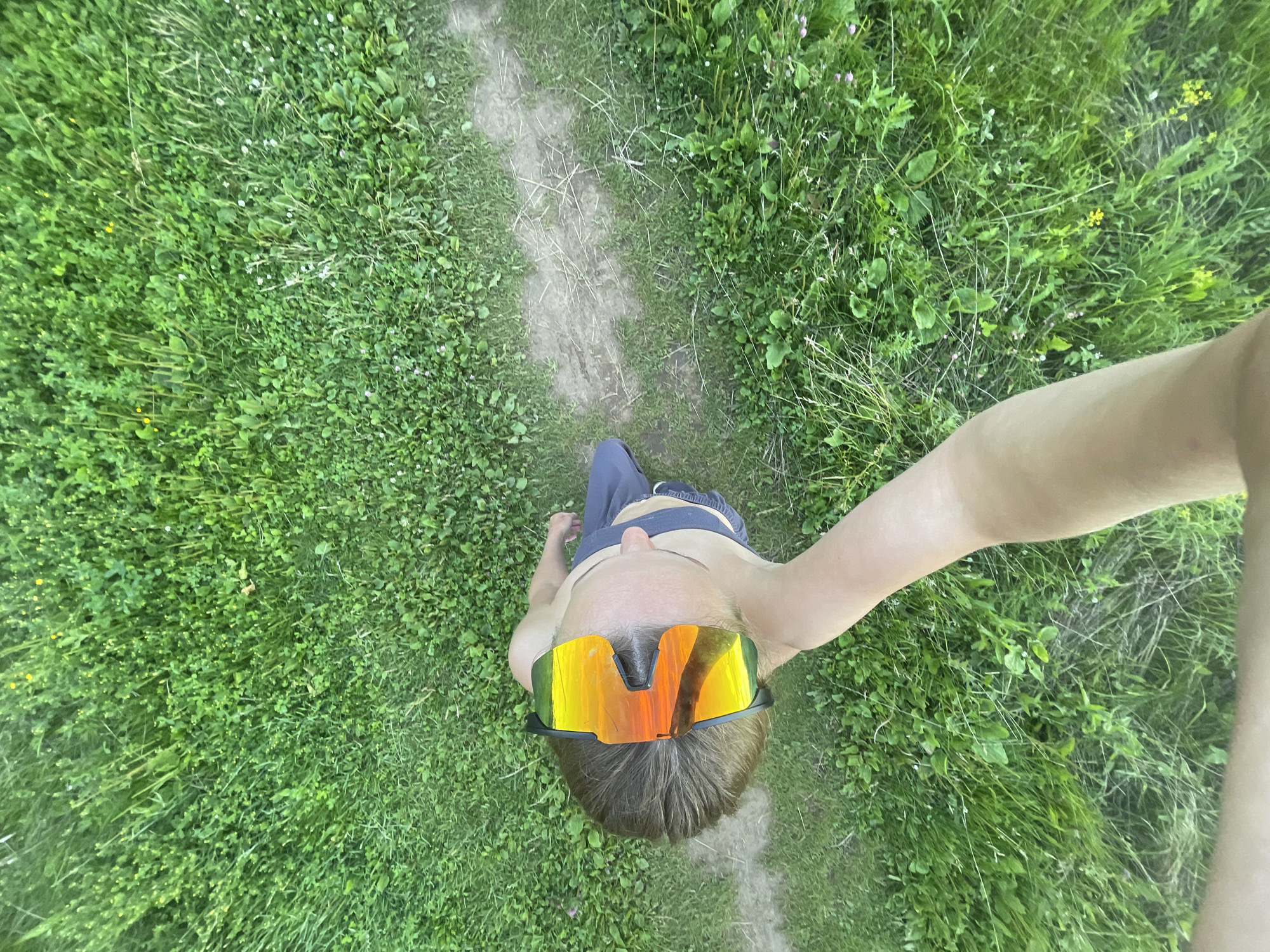 A person is standing on a grassy area with a dirt path running through it. The person is wearing large, orange-tinted sunglasses and dark shorts. The photo is taken from a high angle, looking down at the person. The environment appears to be outdoors, with lush green grass and some small yellow flowers visible. There is no indication of the specific location in the city of Подшивалово, Russia, within the image itself.
