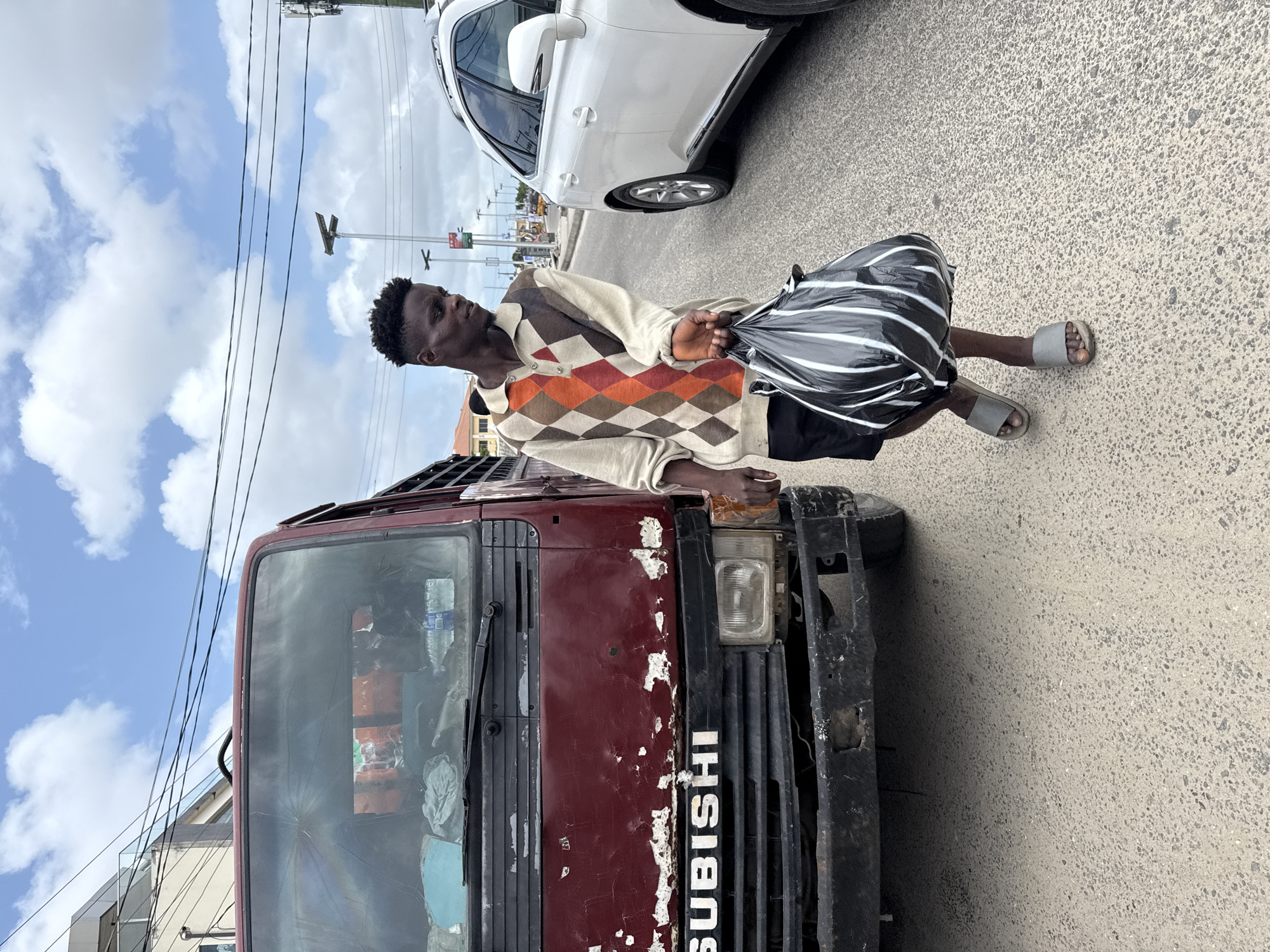 A man stands on the bumper of a maroon Mitsubishi truck on a street in Ado, Nigeria. He is Black, with short hair, wearing a beige and orange argyle sweater, black shorts, and grey sandals, and carrying a black and white striped bag. The truck shows signs of wear with chipped paint and a cracked windshield. A white car is parked nearby on the right. The sky above is blue with scattered white clouds. Power lines are visible against the sky. The scene appears to be on a roadside in an urban setting.