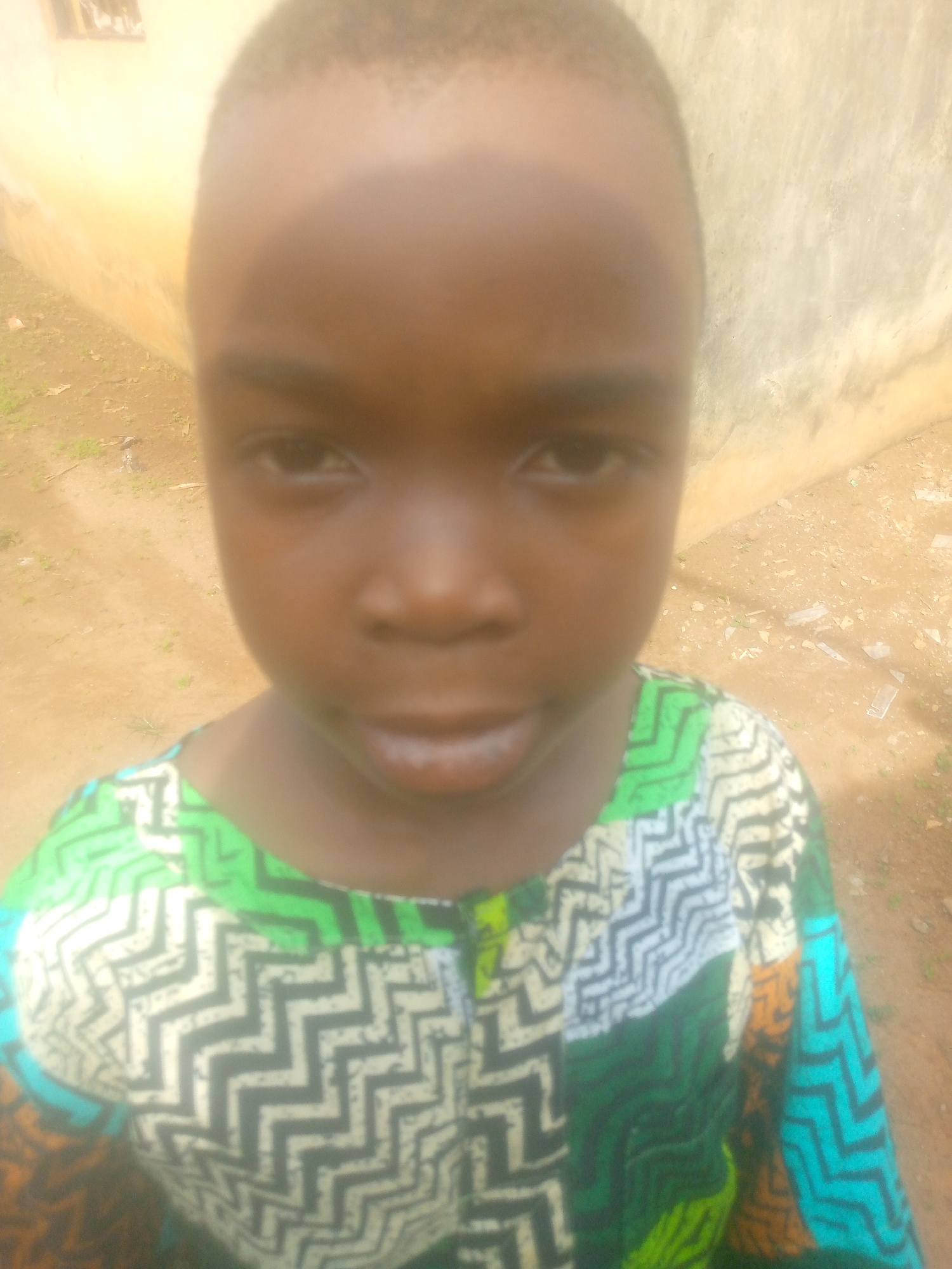 A young Black child stands outdoors, their face the focal point of the image. The child wears a brightly colored shirt with a zigzag pattern featuring green, white, and blue hues. The background consists of a wall and ground with debris, suggesting a residential or public area. The location is Unknown, Unknown. The time of day seems to be daylight, based on the overall brightness.
