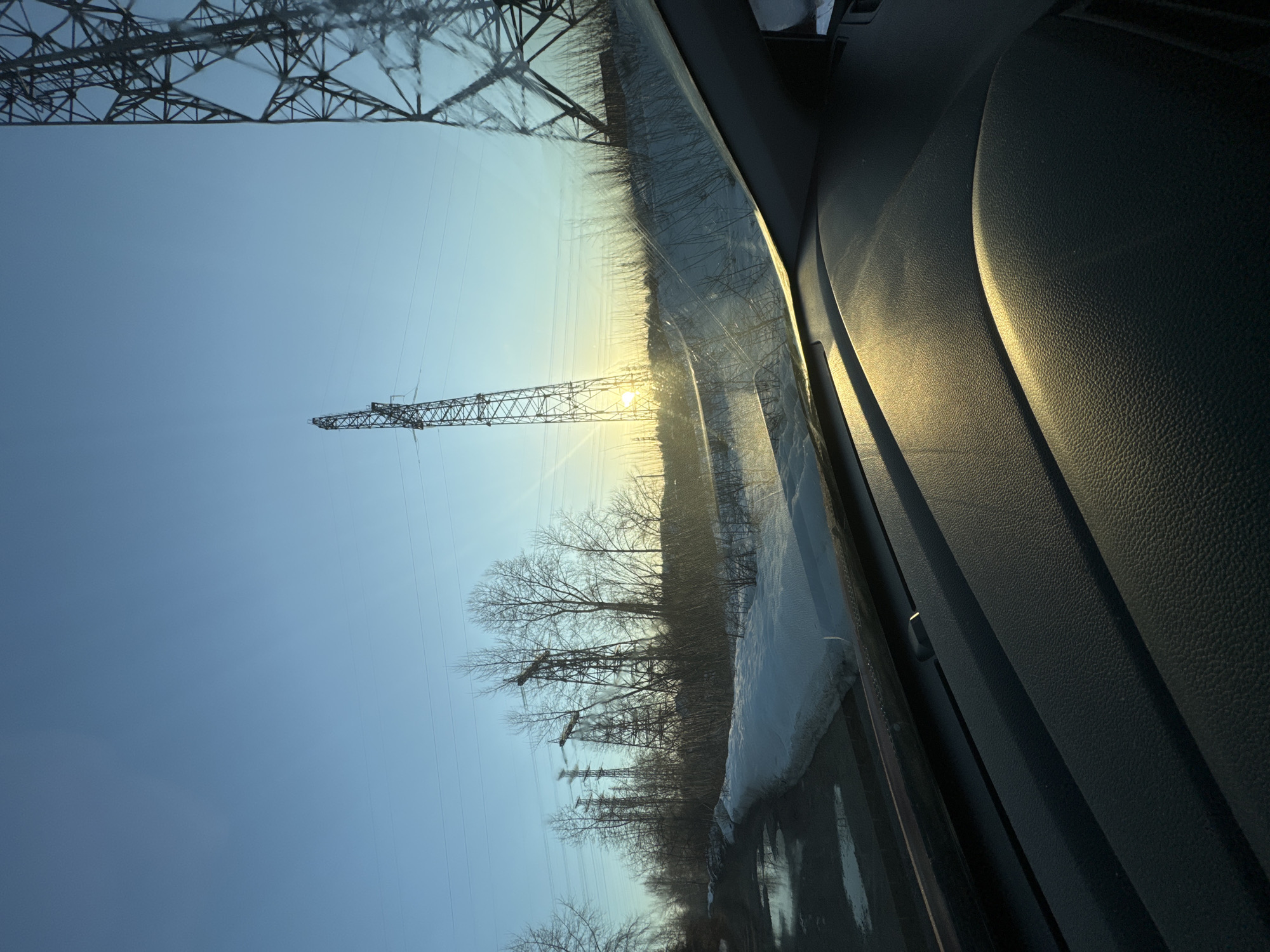 The image, taken from inside a vehicle in what appears to be the Chaikovsky area of Russia, shows a winter landscape with power lines and transmission towers. A layer of snow is visible along the roadside. The sun is low in the sky, possibly indicating early morning or late afternoon. The foreground includes the interior of the vehicle, with a dashboard and the edge of the windshield. No people are visible.