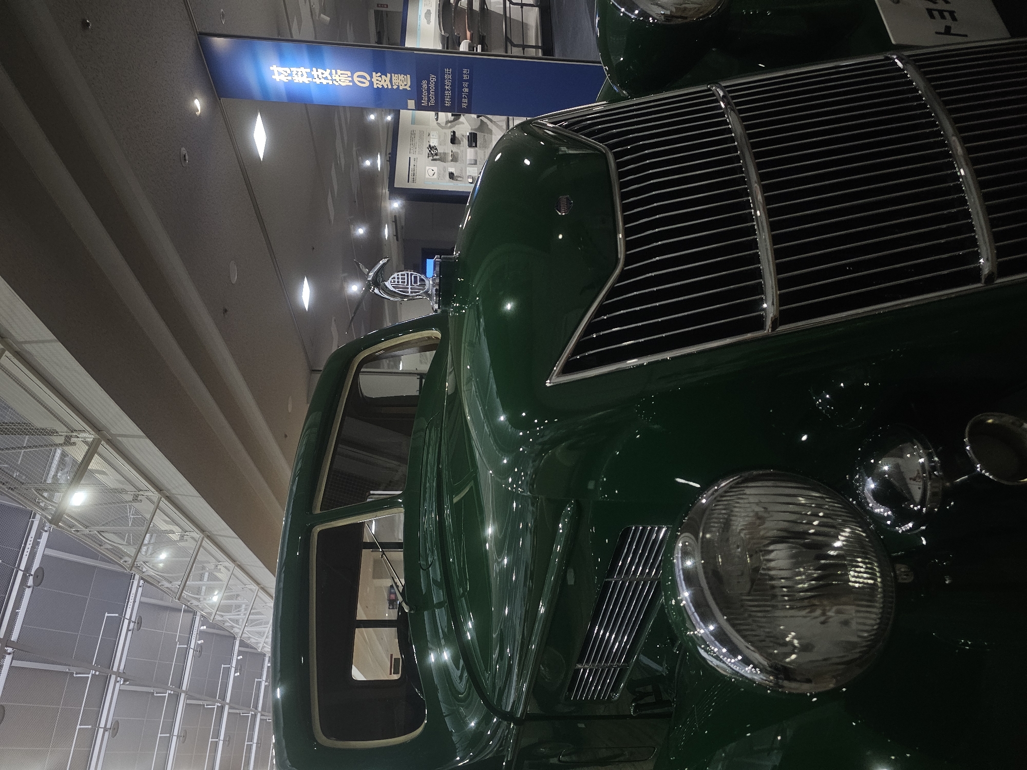 The image shows the front section of a vintage green car parked indoors, likely in a museum. The car features a prominent chrome grille, round headlights, and a hood ornament. The background suggests an exhibition hall with a high ceiling, suspended walkways, and informational displays. One display is a tall blue banner with Japanese text that translates to "Materials Technology." The car appears to be a Toyota Model AA, a model produced in the 1930s, and the overall scene suggests it is on display at the Toyota Commemorative Museum of Industry and Technology in Nagoya, Japan.