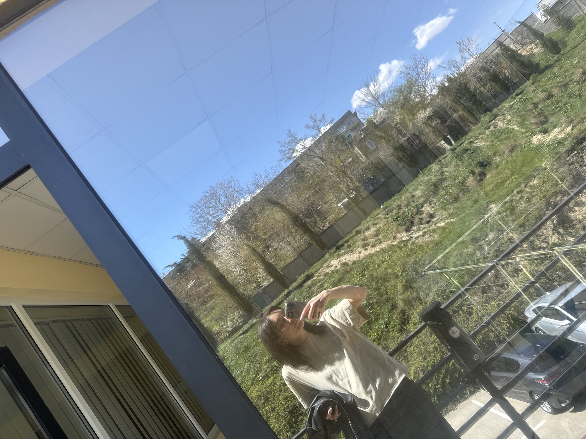 A woman is taking a selfie in front of a large window. The window reflects a cityscape, trees, and green spaces. Cars are visible in the lower right corner, and a fence runs along the edge of the property. The location appears to be Tbilisi, Georgia, based on the architectural style of the buildings in the reflection. The woman is wearing a light-colored t-shirt and dark pants. She is holding a black jacket. The window is part of a building with light yellow walls and white window frames.