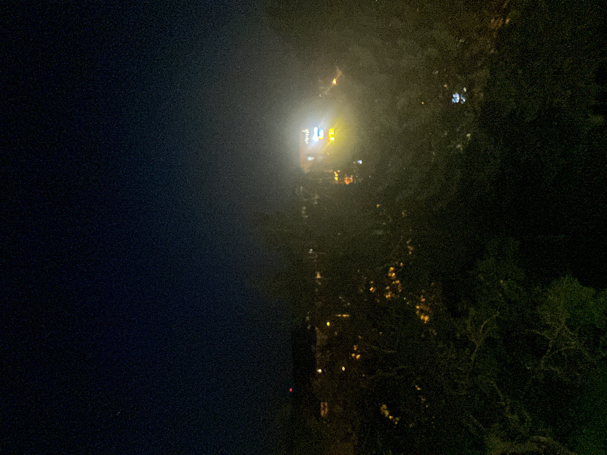 An elevated, slightly blurry nighttime photograph captures a view of a city or town at dusk or night. The sky is dark, with a gradient from deep navy at the top to a lighter gray-blue near the horizon, indicating either twilight or light pollution. In the mid-ground, a cluster of buildings and streetlights are visible, some emitting a warm yellow glow, while others are obscured by atmospheric haze or fog. A particularly bright, rectangular light source, possibly a sign or illuminated building facade, stands out in the center-right of the image, casting a white and yellow glow that is diffused by the mist. In the foreground, dark foliage, likely trees, is silhouetted against the lights, with some scattered yellow lights appearing through the leaves, suggesting more distant illumination. The overall impression is one of a somewhat hazy, urban or suburban landscape at night, with the lights creating a soft, atmospheric effect.