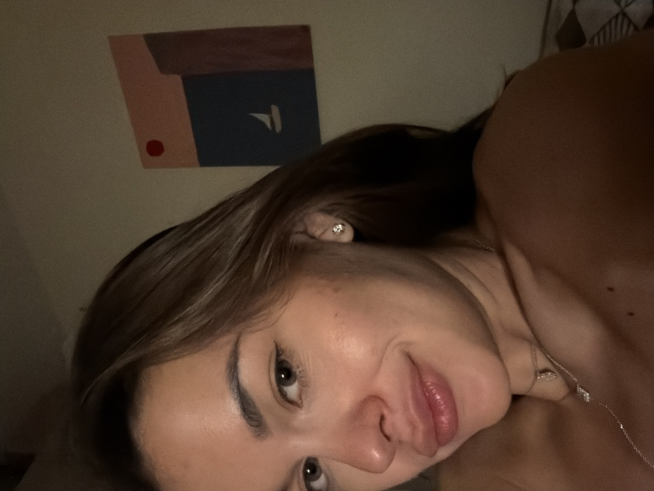 The scene takes place indoors in Tel-Aviv, Israel. The image presents a close-up, mid-torso to head view of a single individual, appearing female, with fair skin and long, medium-brown hair falling over her shoulders. She faces the viewer with a slight, closed-mouth smile. Her eyes are light brown or hazel, and her lips appear full. She wears a small, clear-stone stud earring in her visible right ear and a delicate silver-colored necklace with two small pendants around her neck and upper chest. Her shoulders and upper chest are bare.
The background consists of a plain, light-colored wall. On this wall, a rectangular abstract painting is hung vertically, positioned to the upper right of the individual. The painting features distinct color blocks: an upper section in light peach, a middle horizontal section in dark blue, and a vertical strip on the right in dark reddish-brown. A small red circle is depicted in the upper left of the peach section, and a minimalist white silhouette of a sailboat is centered within the blue section. In the bottom right corner of the image, behind the individual's shoulder, a portion of a light-colored, possibly checkered, patterned fabric is visible. The environment is dimly lit with warm light, suggesting an indoor setting.