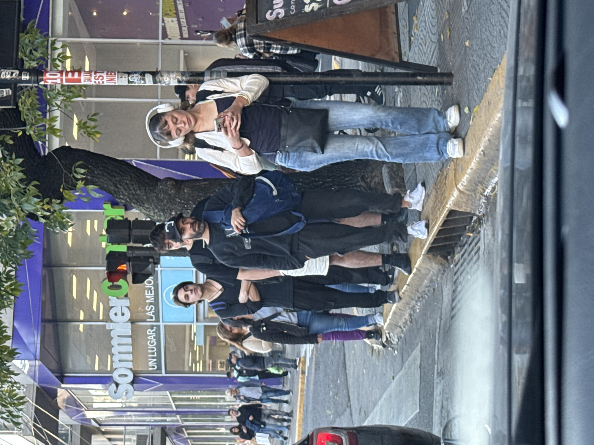 The image depicts a street scene in Buenos Aires, Argentina, with a group of people standing on the sidewalk next to a busy street. The background features a "Sommier Center" store, with the words "UN LUGAR, LAS MEJORES MARCAS" visible.
Several individuals are standing together, with some appearing to be engaged in conversation or waiting. A woman with headphones is looking at her phone. Most of the group is wearing casual clothes like jeans and dark jackets.
The sidewalk is narrow and adjacent to a street with vehicular traffic. A street light and a utility pole are visible along the sidewalk's edge, with a tree trunk and foliage emerging. The overall environment suggests a typical urban setting.