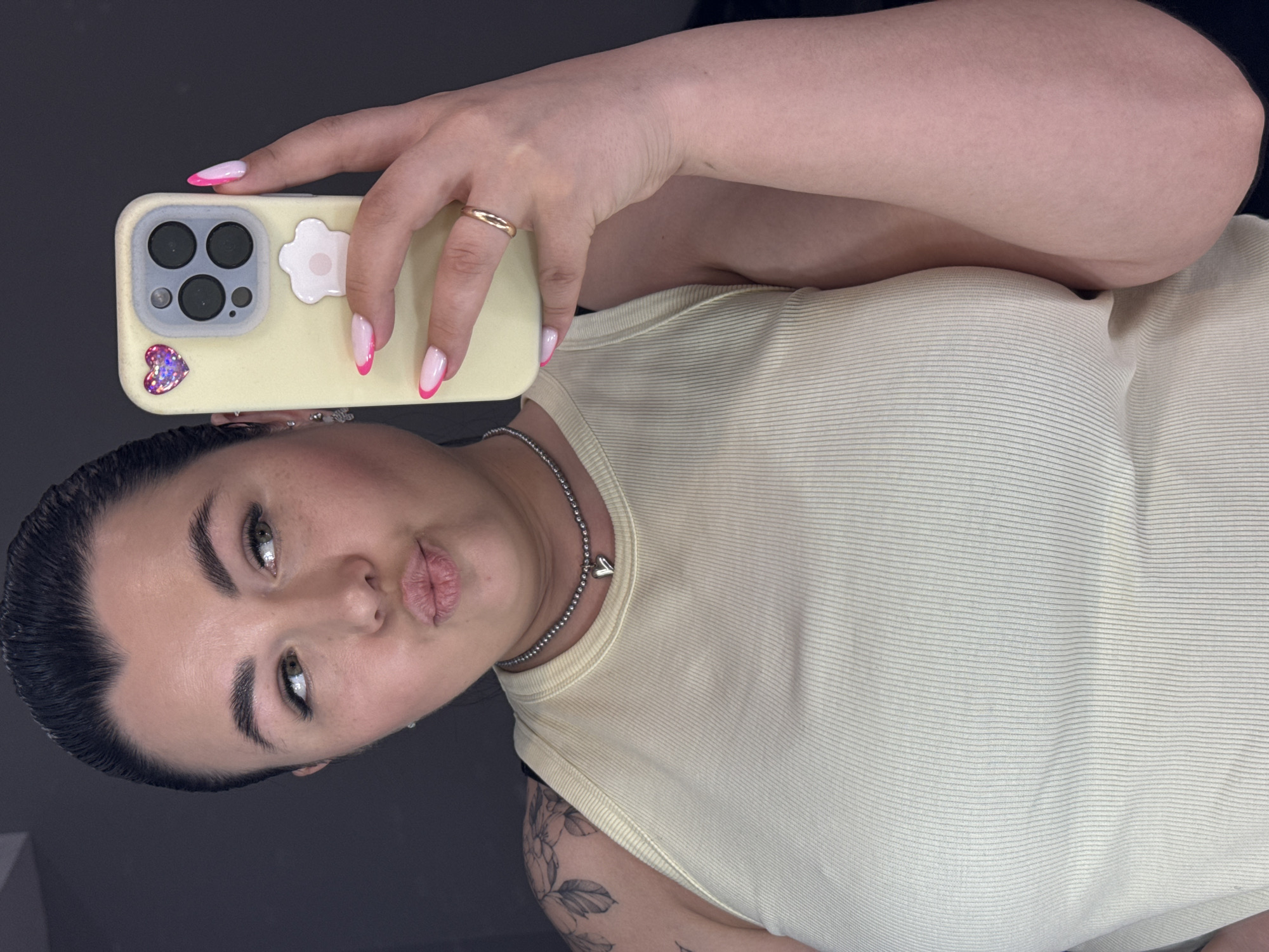 A selfie shows a woman with dark hair pulled back and light skin, wearing a light yellow ribbed tank top and a silver chain necklace with a heart charm. She is holding a yellow iPhone with a pink and white French manicure hand and a gold ring on her ring finger. The phone case has a heart sticker and a flower design on it. A tattoo can be partially seen on her arm. The background is a dark gray or black surface. The location is unknown.