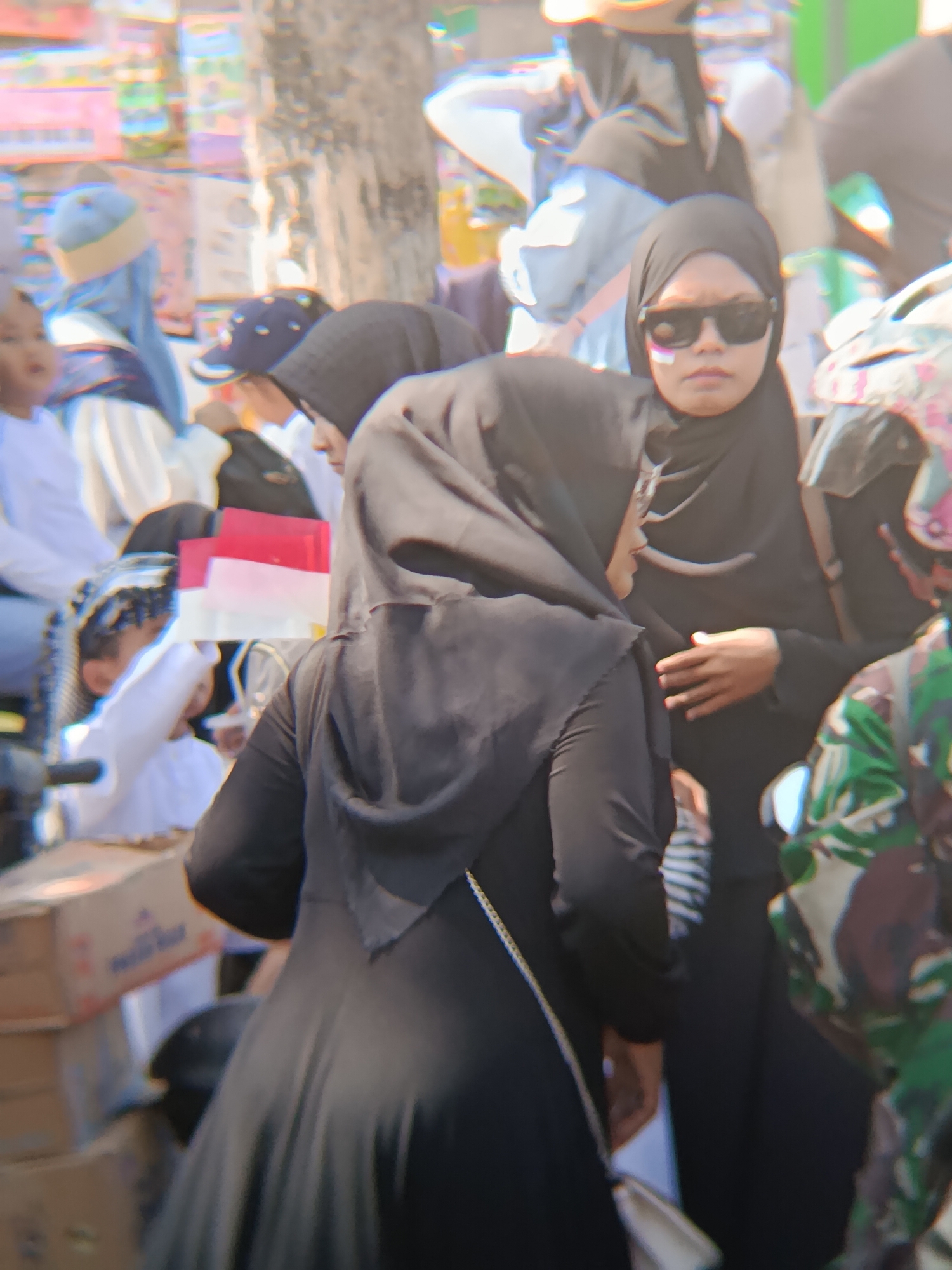 The image depicts a blurred scene of a gathering in Desa Kebon Manis, Kecamatan Cilacap Utara, Indonesia. In the foreground, a person wearing a black hijab and sunglasses is visible from the chest up, facing slightly to the right. They have what appears to be face paint, with a purple and white stripe on their left cheek. To the left of this individual, another person in a black hijab is mostly obscured, seen from the back. In the background, several other individuals are present, some wearing headscarves and one with a baseball cap. There are also some objects, including what seem to be boxes and what could be Indonesian flags (red and white). The environment appears to be outdoors, possibly a street or public area, with a tree trunk visible on the right side of the image. The overall impression is one of a crowded event or celebration, though the details are indistinct due to motion blur and low resolution.
