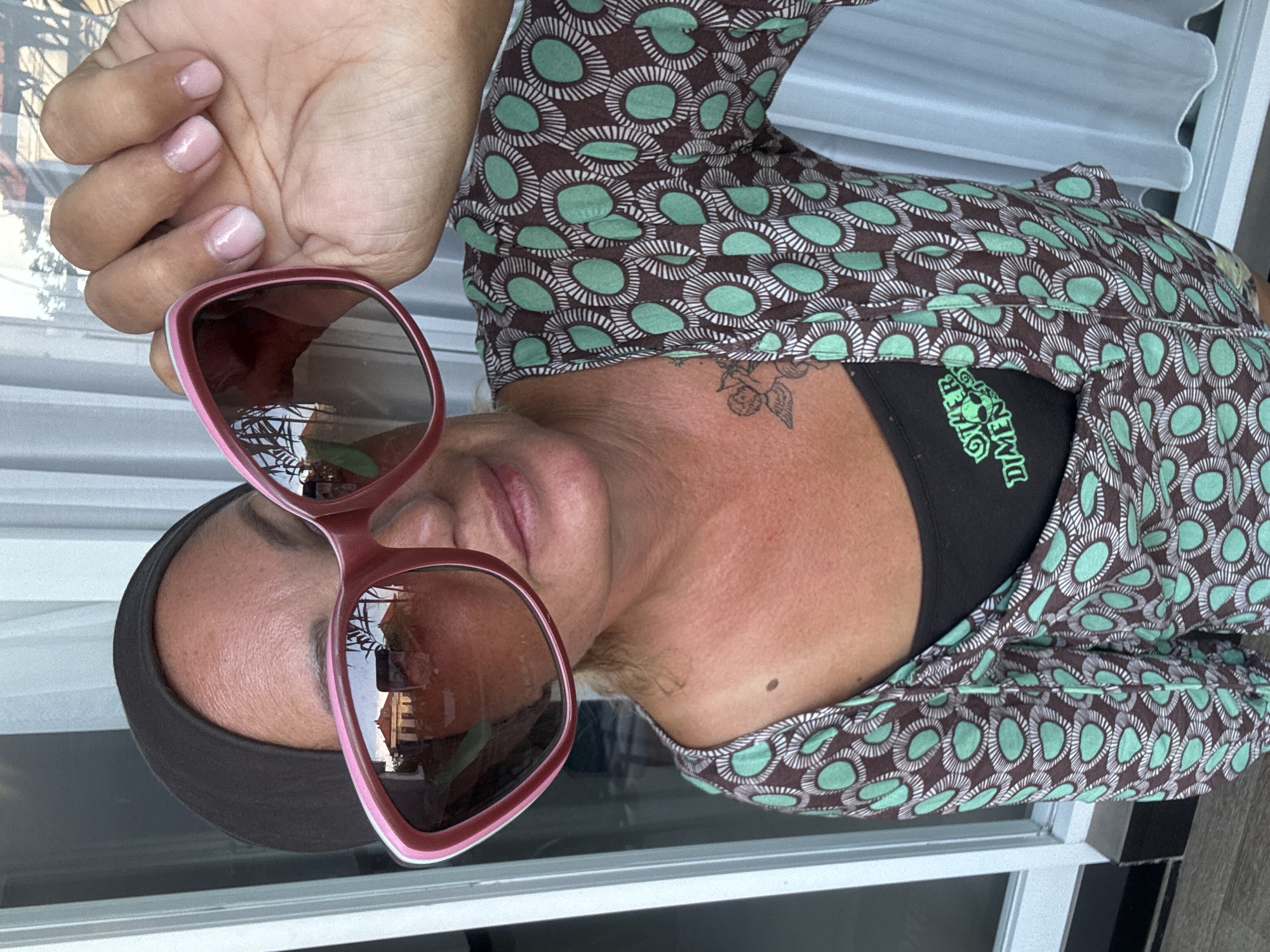 The image presents a medium close-up of a person, rotated 90 degrees counter-clockwise. The individual has tan skin and is wearing a dark brown garment adorned with a repeating pattern of light green and white concentric circles, possibly a top or dress. A black garment, likely a sports bra or athletic top, is visible beneath, featuring stylized white text that partially reads "OVER DIMEN" or "LOVER DIMEN" on the left chest area.
The person is wearing large, square-framed sunglasses with pink rims and dark lenses. The lenses reflect an outdoor scene, including a building with a distinctive red roof, suggesting the person is either outdoors or looking out from a window. A dark cap or headband is worn, covering the forehead. One hand, with light pink nail polish on the fingernails, is visible near the sunglasses.
Other notable details include a small, subtle tattoo resembling a winged figure or cherub on the person's right shoulder or neck area, and a dark mole on the chest. The lighting in the image is bright, indicating it is daytime. The overall setting appears casual, possibly in Kuta, Indonesia, as per the provided context, with a background consisting of white and grey horizontal blinds or louvers. The person's expression is neutral, and the primary activity seems to be posing for the photograph.