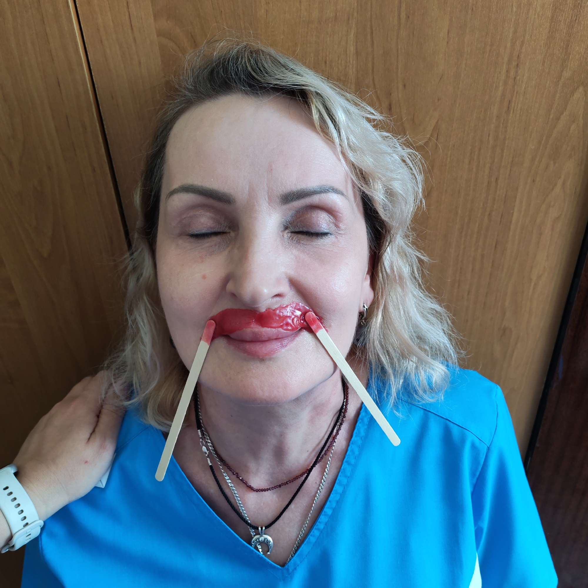 A woman with closed eyes is undergoing upper lip hair removal, as evidenced by the red wax applied to her upper lip and held in place by two wooden applicator sticks. The woman has fair skin, blonde hair, and is wearing multiple necklaces. She is wearing a blue scrubs-like top. Another person's hand rests on her left shoulder. The background consists of wooden panels, likely a door or wall. The location is possibly Chaikovsky, Russia.
