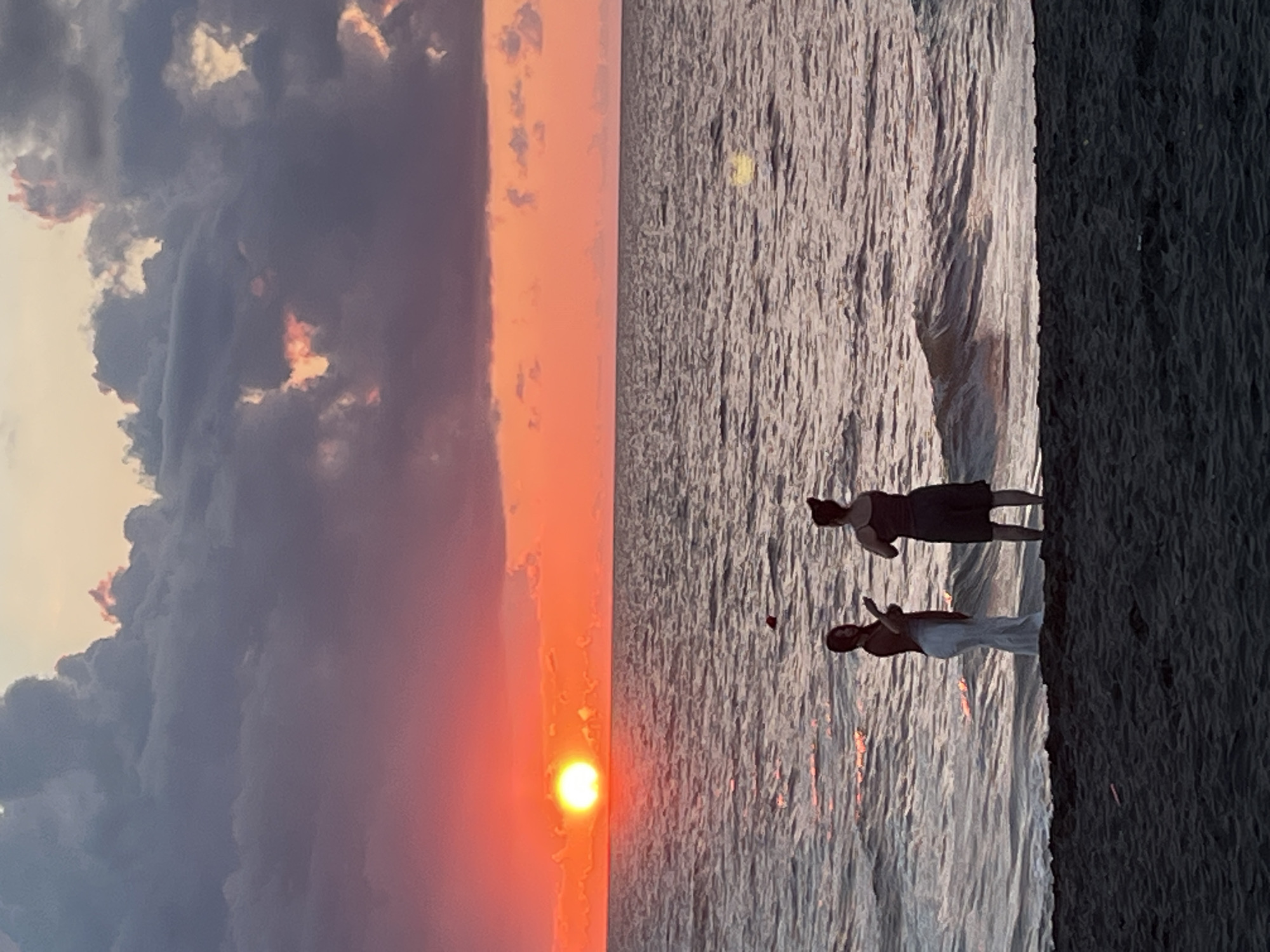 Two individuals are standing on a dark sandy beach in Ureki, Georgia, facing a vast body of water during a vibrant sunset. The sky is a dramatic display of colors, with the sun, a bright orange orb, low on the horizon, casting a warm glow across the water. Dark, heavy clouds are interspersed with patches of pink and orange, creating a moody yet beautiful atmosphere.
The person on the left, appearing to be female, is wearing a long white garment and standing closer to the water's edge. The person on the right, also appearing to be female, is wearing dark shorts and a dark top, standing slightly further back on the beach. Both individuals are silhouetted against the bright sky, their features obscured. They appear to be observing the sunset, possibly taking pictures or simply enjoying the view.
The water is choppy, with small waves rolling towards the shore, reflecting the colors of the sunset. The beach itself is dark and appears to be composed of fine sand. The overall scene is one of natural beauty and tranquility, with the human presence adding a sense of scale and narrative to the picturesque setting.