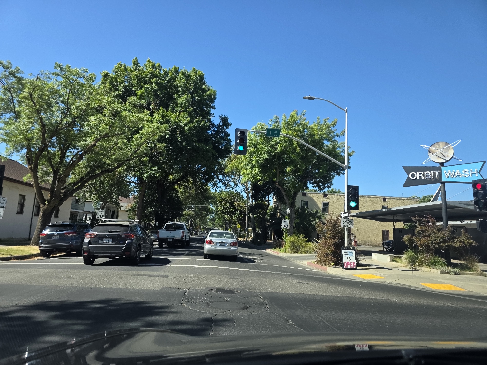 This media file captures a bright, sunny day in Sacramento, California, likely during midday given the strong shadows and clear blue sky. The scene is a street intersection with multiple cars, trees lining the road, and buildings in the background.
In the foreground, the perspective is from inside a vehicle, showing the dashboard and windshield. Several cars are visible on the road. A dark SUV with license plate AXOR076 is in the left lane, while a lighter-colored sedan is ahead in the right lane. Further down the road, a white pickup truck is also visible.
Traffic signals are prominent, with a green light illuminated, indicating traffic can proceed. A street sign indicates "T ST" and another reads "SPEED LIMIT 30". To the right, a sign for "ORBIT WASH" with a circular, atomic-like logo is clearly visible, along with a red traffic signal. A sign on the sidewalk states "IS OPEN".
The presence of mature trees suggests a well-established neighborhood. The overall impression is a typical urban street scene on a clear, warm day. There are no people clearly visible in the image. The focus is on the road, vehicles, and surrounding infrastructure.