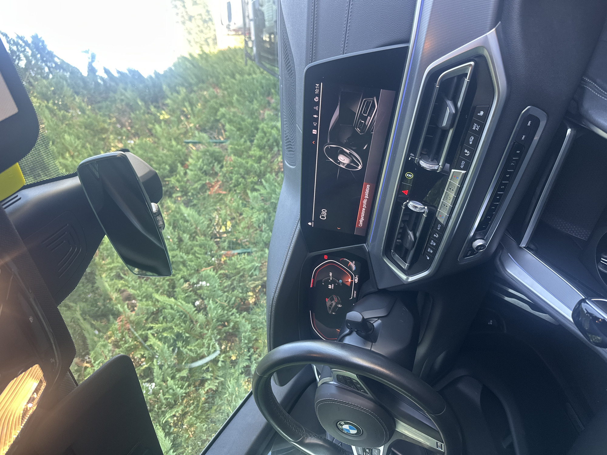 The image shows the interior of a modern car, likely a BMW, parked outdoors. The viewpoint is from the driver's seat, looking towards the dashboard and windshield. The car's dashboard features a large, central touchscreen display showing a greeting "Ciao" and a graphic of the car's interior, along with navigation information. To the left of the screen is the steering wheel, which has a BMW logo in the center. The instrument cluster behind the steering wheel is also visible, displaying vehicle information. The car's interior is dark, with leather accents. Outside the car, through the windshield, lush green trees and vegetation are visible, suggesting a natural or park-like setting. The sky is bright, indicating daylight. The specific location in Fornoli, Italy, cannot be determined from the image alone.