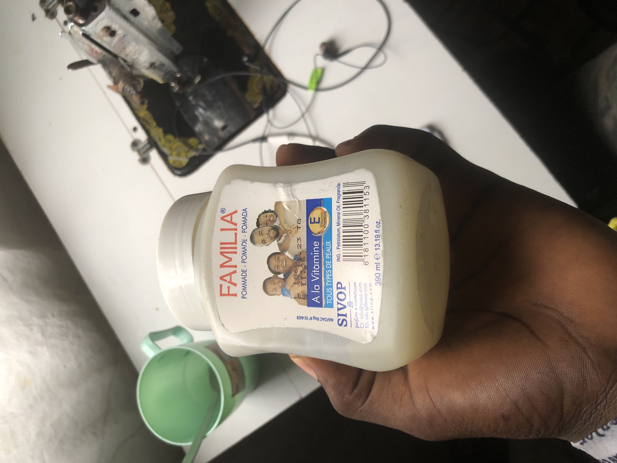 A person's hand is holding a white plastic jar of SIVOP hair pomade, which has a label with a picture of a family. The background shows a white table with a green plastic cup and part of a sewing machine. The image appears to be taken in a home environment in Ado, Nigeria.