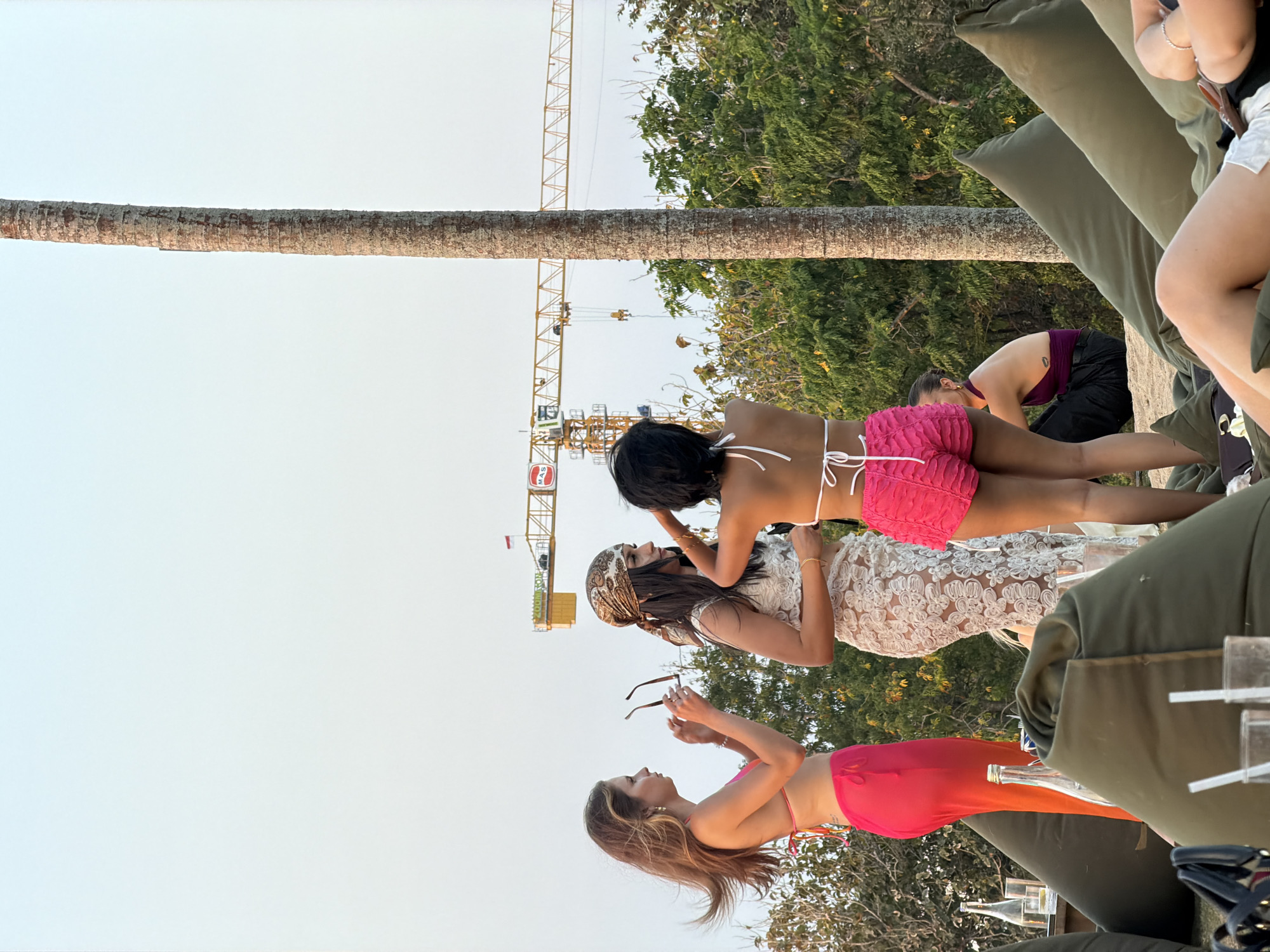 Four women and one person are visible outdoors. The scene appears to be a beach or resort setting with sand, palm trees, and lush green foliage in the background. A yellow construction crane is also visible in the distance.
In the foreground, three women are standing and interacting. The woman on the left has long brown hair and is wearing a pink bikini top and a sheer orange skirt. She is holding a pair of sunglasses. The woman in the middle has dark hair covered by a patterned scarf and is wearing a white lace dress. She is looking to the right. The woman on the right has short dark hair and is wearing a white bikini top and pink ruffled shorts. She is facing away from the camera.
In the mid-ground, a person wearing a dark purple top and black pants is seated, facing towards the right. They have a tattoo on their left shoulder blade. Several large, olive-green beanbag chairs are scattered throughout the scene, with some individuals seated on them in the background. Bottles and glasses are visible on a surface near the beanbag chairs. The sky is a pale blue with a slight gradient towards white at the top.