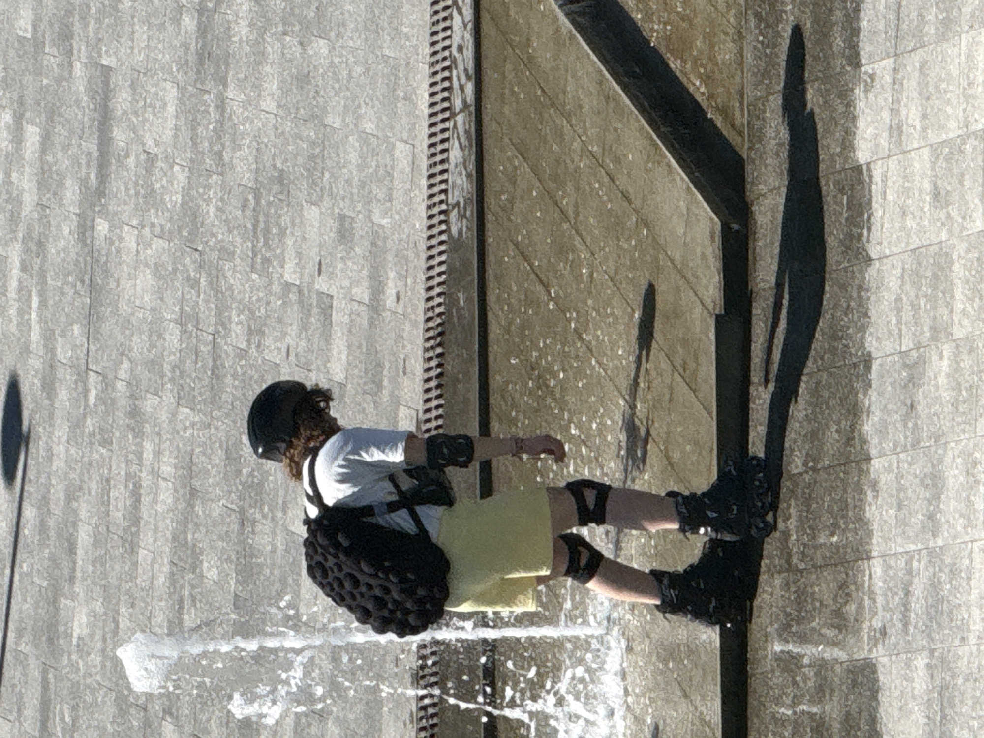 A person wearing a helmet, black backpack, light-colored t-shirt, yellow shorts, knee pads, elbow pads, and roller skates is partially visible, bending forward near a water fountain. The person's face is not visible. The ground appears to be paved with rectangular stones, with some areas wet from the fountain. A dark shadow is cast on the ground to the right of the person.