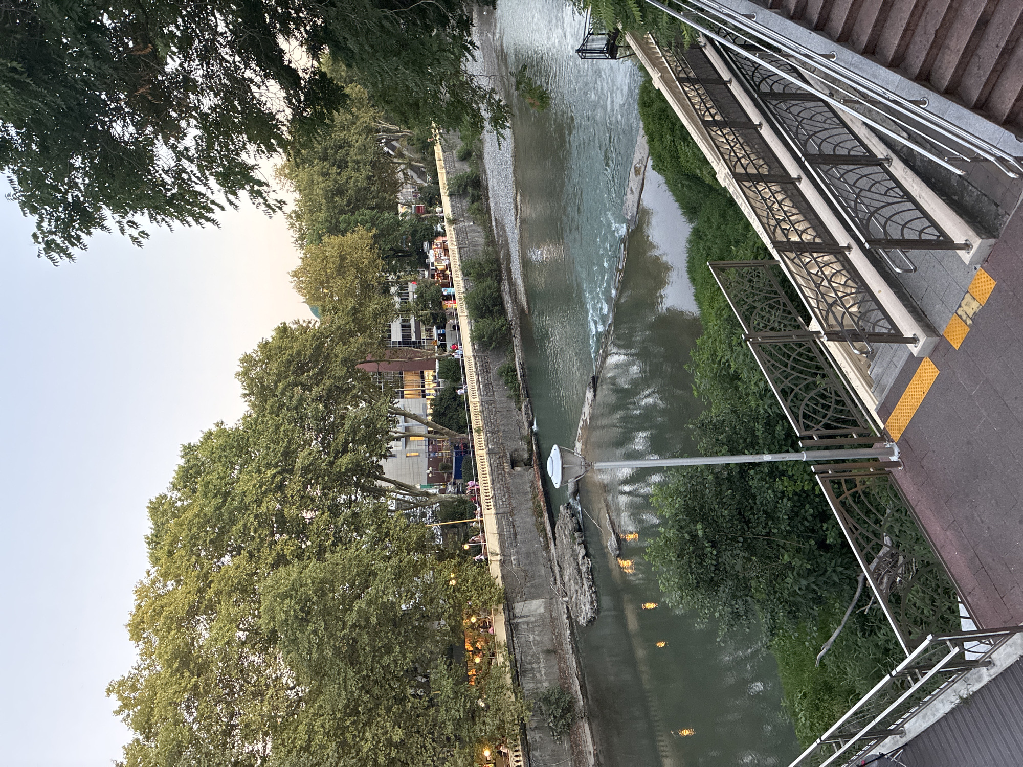 The image depicts a view of the Mzimta River in Sochi, Russia, near the city center. A pedestrian walkway with ornate metal railings runs alongside the river, with a set of stairs descending from the foreground to a lower level. The river flows gently, with some visible rapids and scattered rocks in the water. On the opposite bank, a stone retaining wall supports a street lined with trees, buildings, and streetlights. Several people are visible walking along the street and on the retaining wall. The sky is clear and appears to be late afternoon or early evening. The overall atmosphere is one of urban tranquility and natural beauty.