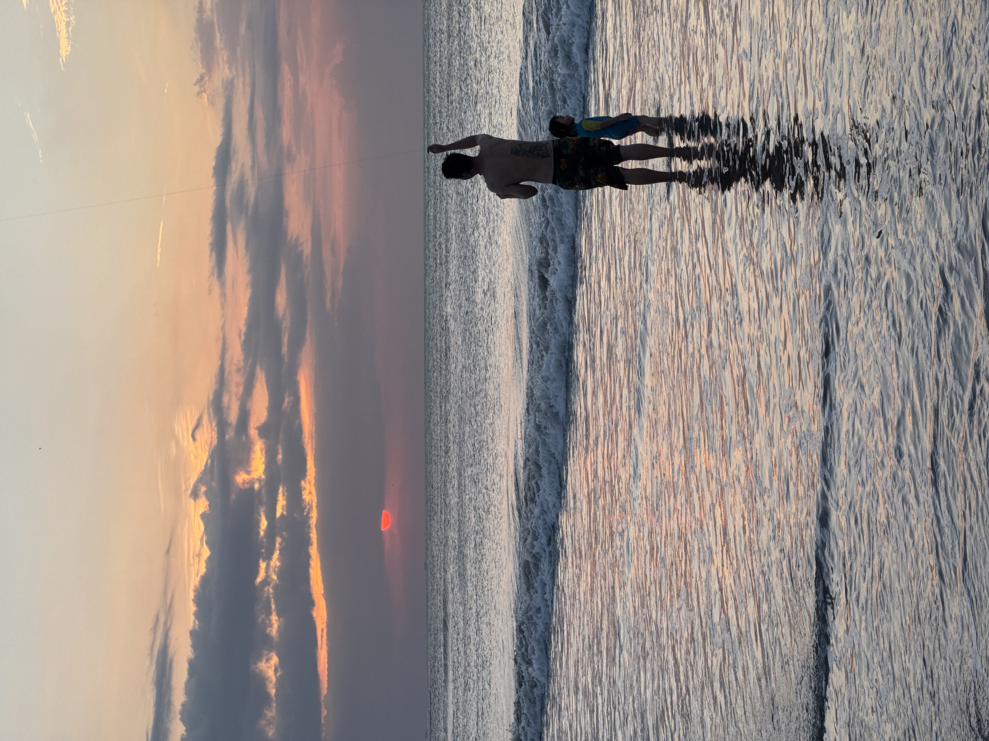 The image depicts a father and child standing in shallow ocean water, with waves gently lapping around their legs. The father, shirtless and wearing patterned swim trunks, holds his arm up, seemingly flying a kite, as a thin line stretches upwards into the sky. Beside him, the child, wearing a blue and yellow swimsuit, stands facing the ocean. The sun is setting over the horizon, casting a warm, orange and pink glow across the sky and reflecting on the water's surface. Dark clouds are present in the upper portion of the sky, with some breaks revealing the sunset colors. The scene appears to be a tranquil evening at a beach. Based on the tropical setting and likely tourist activity, a plausible location is Kuta Selatan, Indonesia, known for its popular beaches.