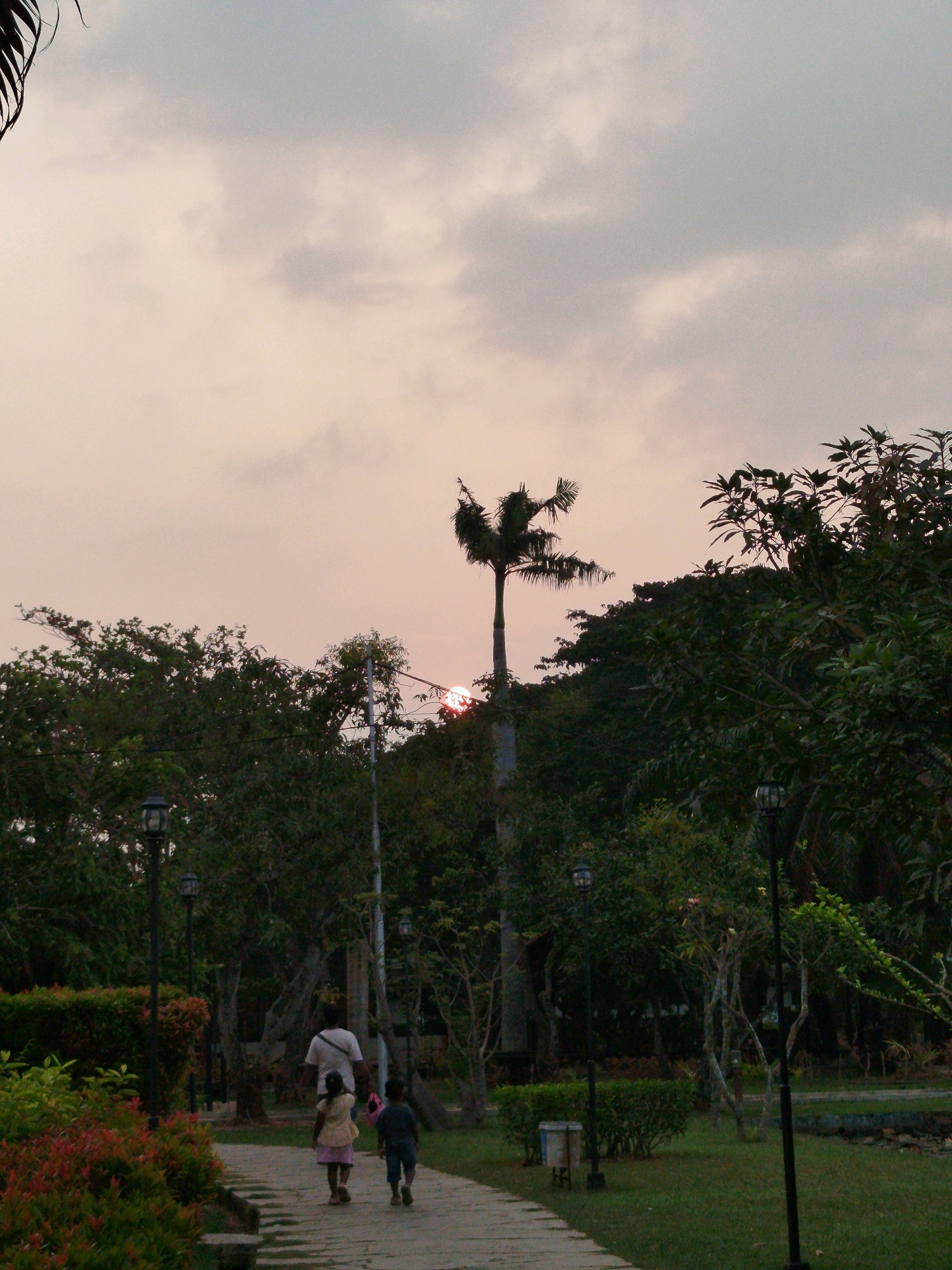 The image depicts an outdoor scene at dusk in Bugangin, Indonesia. A paved pathway meanders through a park-like setting, bordered by lush green grass and dense foliage. Several street lamps are visible, casting a soft glow. In the foreground, a man walks with two children on the pathway. The man is wearing a white t-shirt and dark pants, while the girl wears a yellow dress and the boy is dressed in a blue t-shirt and shorts. The background features trees and a palm tree, with a faint glow in the sky suggesting the setting sun. The overall atmosphere is tranquil.