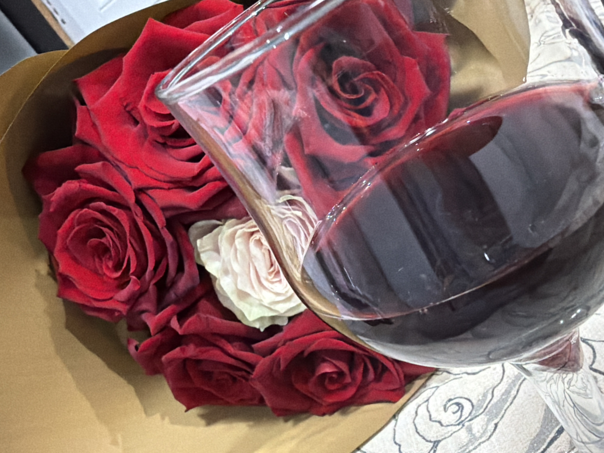 An intimate close-up shot captures a bouquet of roses and a wine glass filled with red wine, suggesting a romantic or celebratory occasion. The bouquet, predominantly composed of deep red roses, is artfully wrapped in brown kraft paper, occupying the left and central portions of the frame. One lighter-colored, possibly white or pale pink, rose is nestled among the vibrant red blooms, adding a subtle contrast.
In the foreground, a clear wine glass is tilted, holding a generous pour of dark red wine. The glass reflects light, and its transparent bowl allows a distorted yet detailed view of the roses behind it. The wine itself is a rich, dark hue.
The scene is set indoors, likely on a table or flat surface, partially covered by a light-colored, patterned textile visible in the bottom right corner and extending under the wine glass. This textile features abstract or floral patterns. The lighting appears soft and artificial, characteristic of an evening or indoor setting, with no direct sunlight. No people are visible, and there is no discernible text within the image. The overall impression conveys a sense of warmth, elegance, and a special moment, potentially taking place in Pazardzhik, Bulgaria, though no specific local cues are present in the visual.