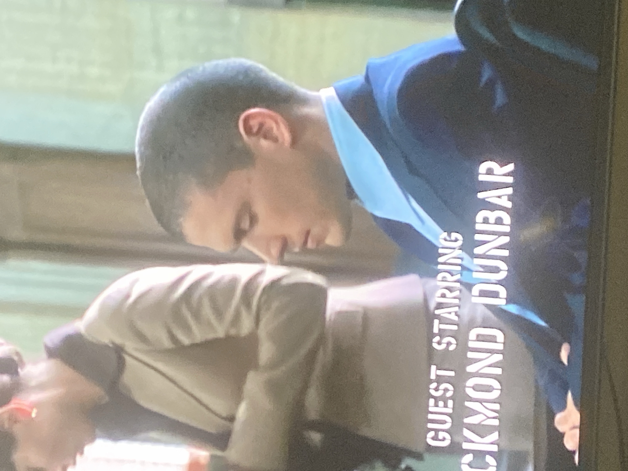 This image, likely a still from a film or television show, shows two men partially visible in an indoor setting. The scene appears to be taken from a screen, featuring overlaid text credits.
In the foreground (bottom of the rotated image), a man is shown bent forward, wearing a light-colored jacket over a darker garment. His head is visible, and he appears to be looking downwards. He may be wearing glasses, but it's not entirely clear due to the image quality.
In the background (top of the rotated image), another man is also bent forward, wearing a dark blue suit jacket over a light blue collared shirt. He too is looking downwards, with his head and upper torso visible. Both men's posture suggests they are observing something below them or engaging in an activity that requires them to lean forward.
The background is softly blurred with indistinct green and brown tones, consistent with an indoor environment or a staged set. The lighting is diffused, and no specific time of day can be determined.
Prominently displayed over the image, vertically aligned, are white text credits. The top line clearly reads "GUEST STARRING". Below this, the partial name "CKMOND DUNBAR" is visible. Given common actor names, this likely refers to "ROCKMOND DUNBAR".
While the location context is provided as Gosa, Nigeria, there are no specific visual cues within the image itself to confirm this geographical location. The scene appears to be a generic interior. The emotions of the subjects are not discernible due to their posture and the focus on the credits.