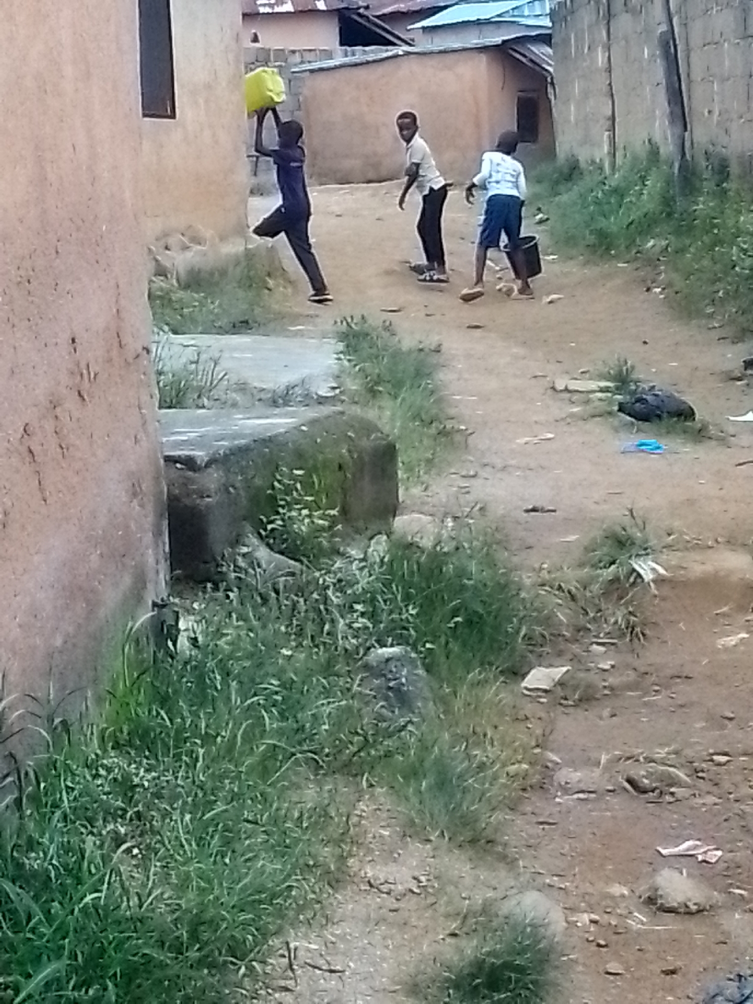 In an outdoor, urban environment, likely a village or informal settlement, three young individuals are depicted on a dirt path. On the left, a person is shown in mid-air, jumping with a bright yellow container held aloft in their right hand. This individual is wearing dark clothing, including a long-sleeved shirt and pants, and dark shoes. To their right and further down the path, two other individuals are visible. One, wearing a light-colored shirt and dark pants, is facing the camera. Next to them, another person, dressed in a light-colored long-sleeved shirt and blue shorts, is seen from behind, holding a black bucket. The ground is predominantly dirt, with scattered rocks and sparse green vegetation lining the path. Buildings with muted-colored walls and corrugated metal roofing are present in the background. The overall scene suggests a moment of activity, possibly play or a chore, in a modest, undeveloped area.