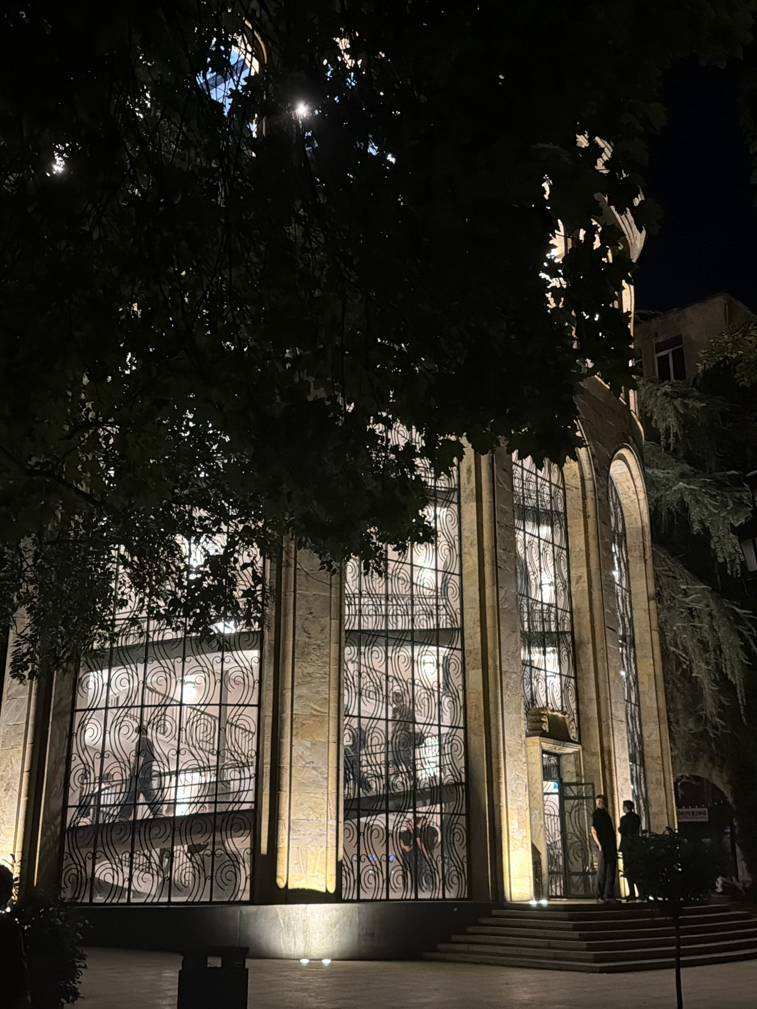 The image captures the exterior of the National Parliamentary Library of Georgia in Tbilisi at night. The building features large, arched windows with intricate black metal grills. Warm, yellow light illuminates the facade from spotlights at the base of the building, creating a dramatic effect. Silhouettes of people can be seen through the windows, suggesting activity inside. Two individuals stand on the steps leading up to the entrance, appearing to converse. The foreground is a paved area, possibly a plaza, with a trash receptacle visible. Lush trees with dark foliage frame the scene, their branches partially obscuring the view of the building and the night sky, where the moon is faintly visible through the leaves. The overall atmosphere is one of quiet evening activity in a historic urban setting.