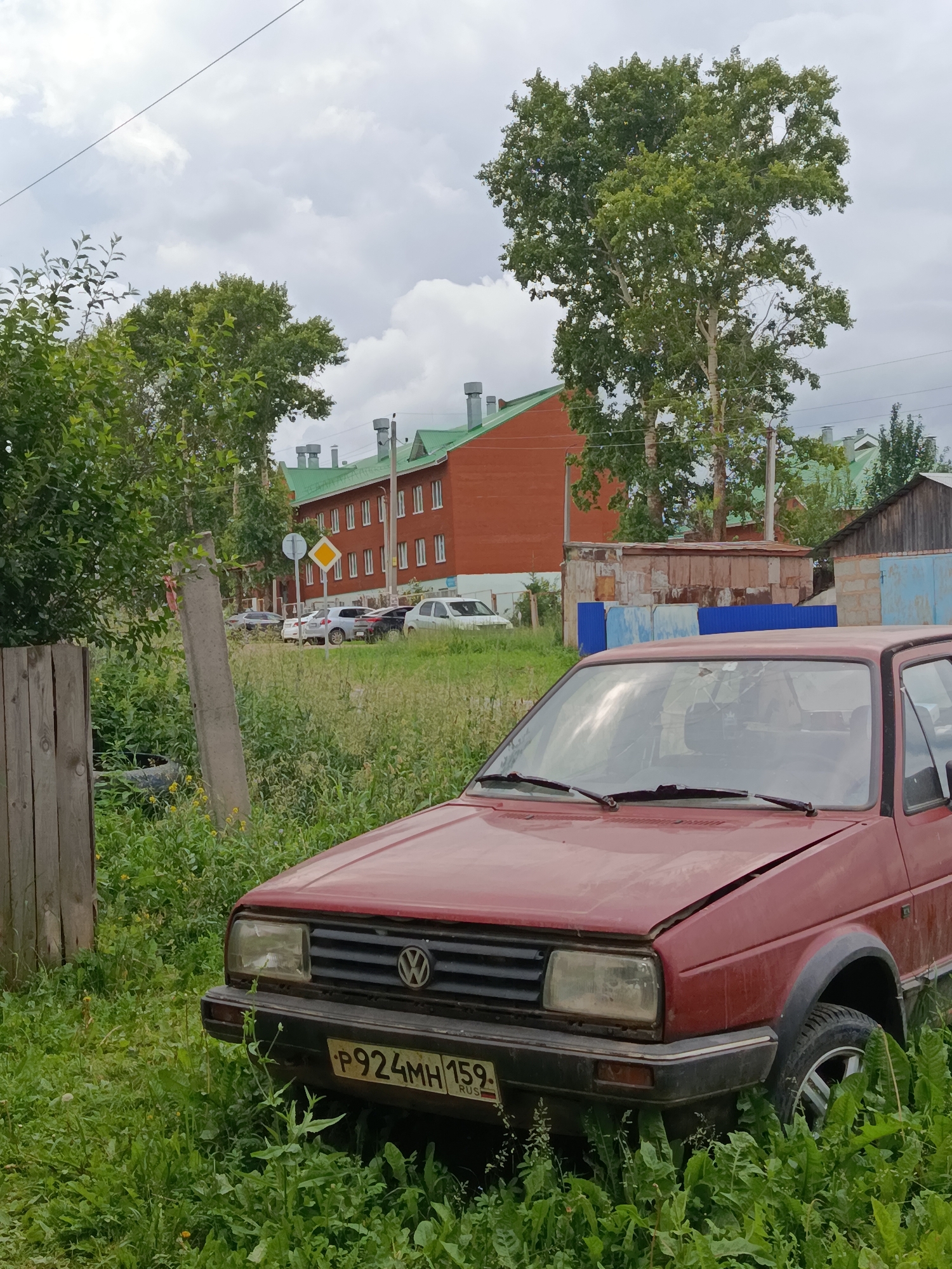 The image captures a weathered, red Volkswagen hatchback parked in a grassy area. The car appears to be old and possibly abandoned, with its hood slightly ajar and the paint showing signs of age. The license plate is visible and reads "P924MH 159 RUS". In the background, a multi-story brick building with a green roof and a row of identical windows suggests an urban or semi-urban setting. Several other cars are parked in front of this building. Further back, more buildings with green roofs are visible, along with trees and utility poles. A wooden fence post and some scattered debris are present in the foreground. The sky is overcast with clouds. The overall scene depicts a somewhat unkempt environment, likely a residential or industrial area within the city of Fokino, Russia, given the license plate.