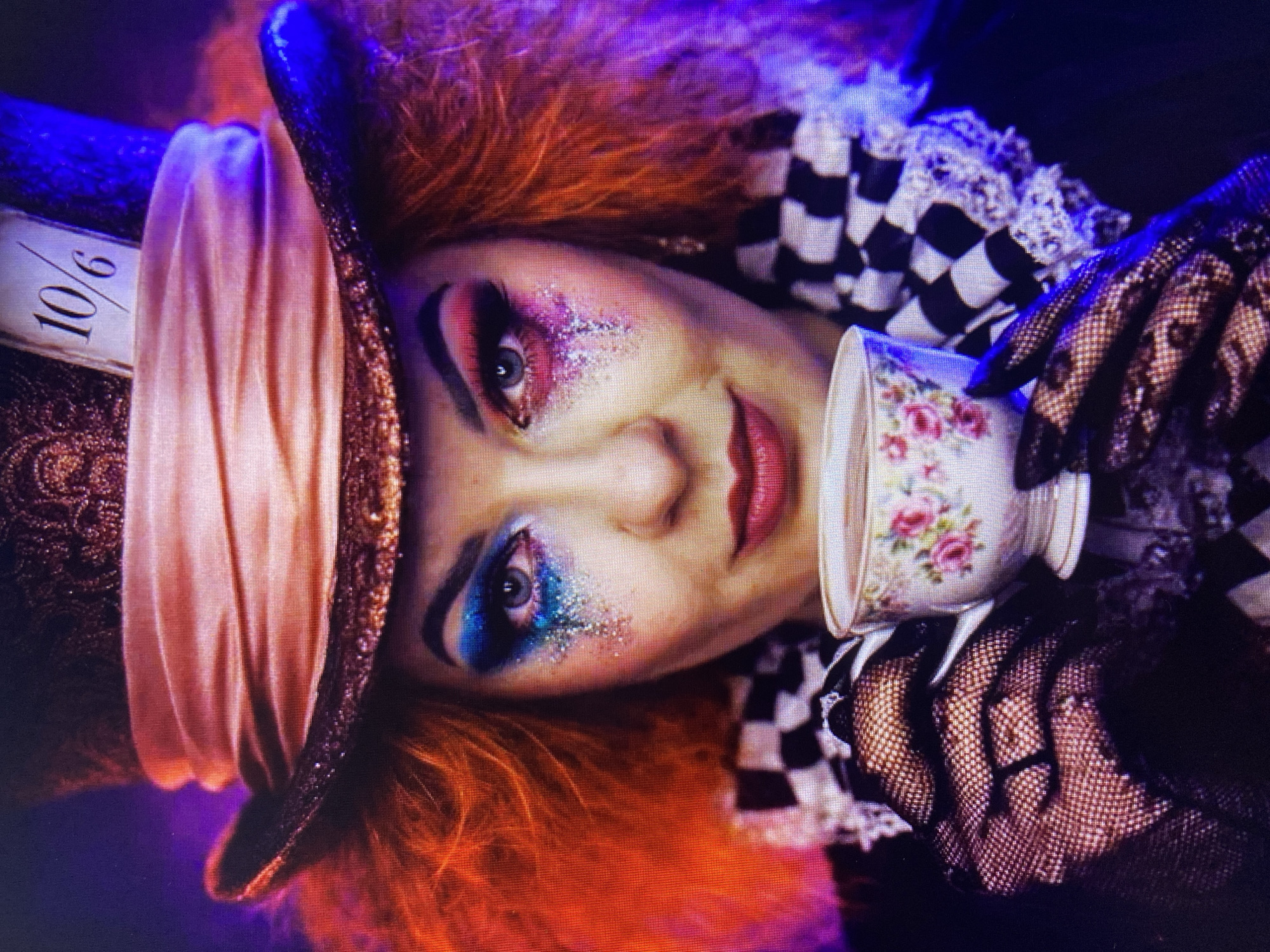 A person in elaborate makeup and costume, resembling the Mad Hatter from Alice in Wonderland, holds a teacup. The individual has vibrant red hair, dramatic blue and pink eyeshadow with glitter, and is wearing a large, ornate hat with a "10/6" tag. They are also dressed in a black and white checkered collar with lace trim and black fishnet gloves. The background is dark with purple and blue lighting. The image does not contain any information about the city of Stavropol, Russia.