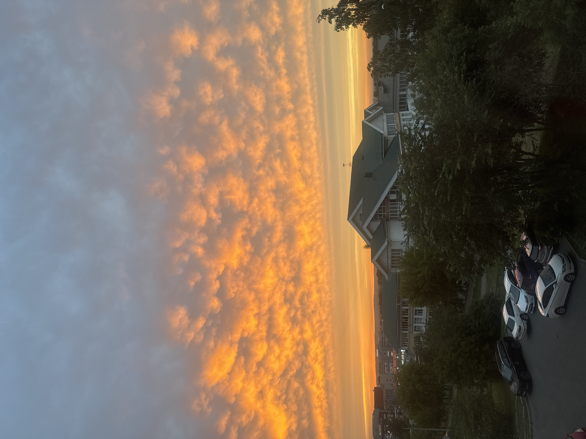 The image depicts a dramatic sunset over a city landscape. The sky is dominated by vibrant orange and yellow clouds, forming a textured pattern that stretches across the upper portion of the frame. Below the sky, a low-rise building with a white facade and green roof is visible, set against a backdrop of trees. In the foreground, several parked cars are arranged on a paved surface, suggesting a residential or commercial area. The overall scene captures the atmospheric beauty of a sunset in an urban environment. Given the context of Irkutsk, Russia, the image likely represents a view from an elevated position within the city, possibly from an apartment building or a higher floor of a structure. The architecture of the building and the presence of trees are consistent with urban settings in Russia.