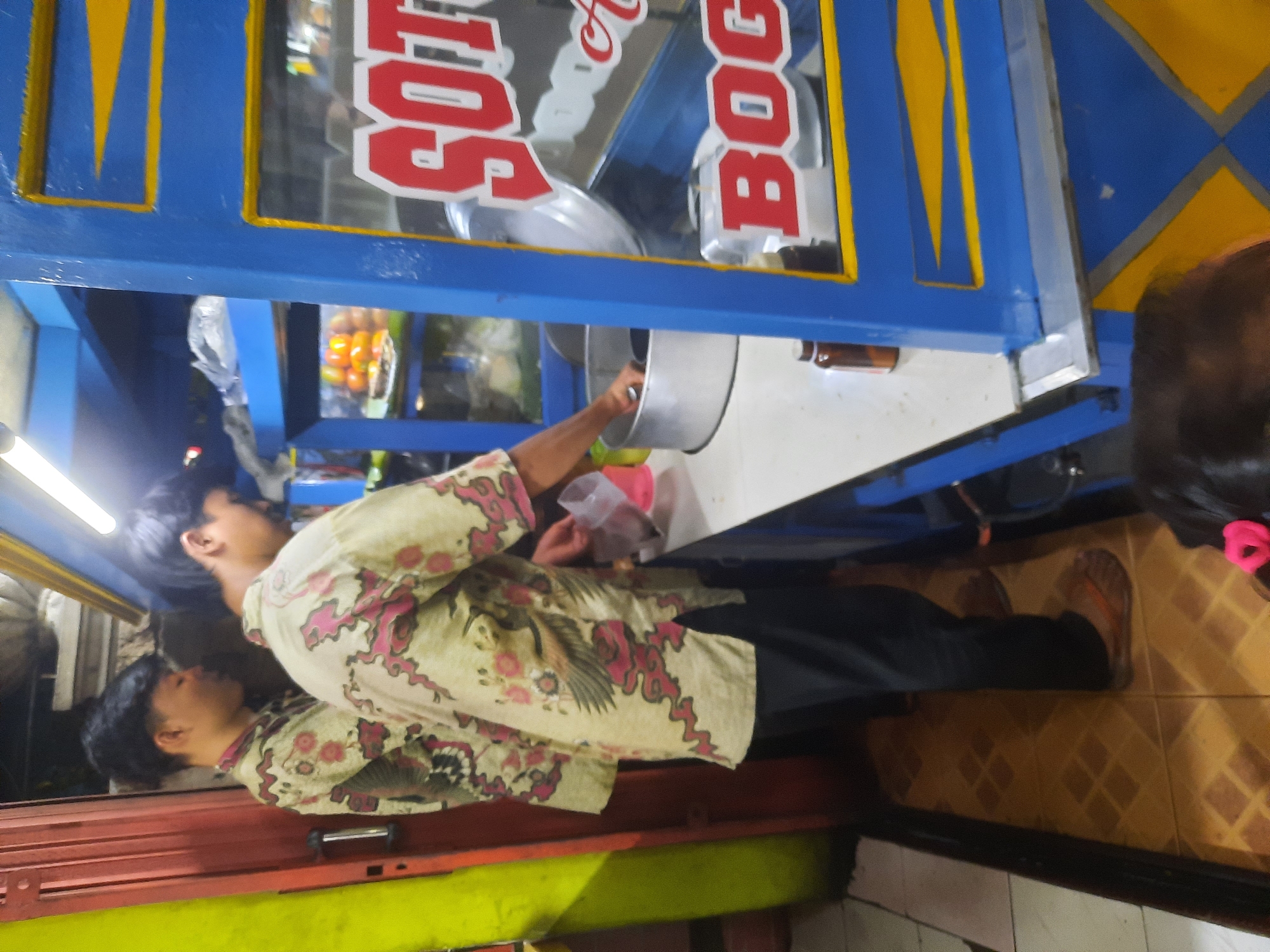 Two young men, likely employees, are working at a food stall in North Jakarta, Indonesia. The stall is brightly decorated with blue and yellow paint and features prominent red lettering that appears to spell out "SOTO BOGOR." One man, wearing a patterned shirt and dark pants, is actively engaged in preparing food, reaching into a large metal pot. The other man, also in a similar patterned shirt, stands beside him. Behind them, shelves display food items, including oranges, and a light fixture illuminates the area. The floor is tiled with a diamond pattern.