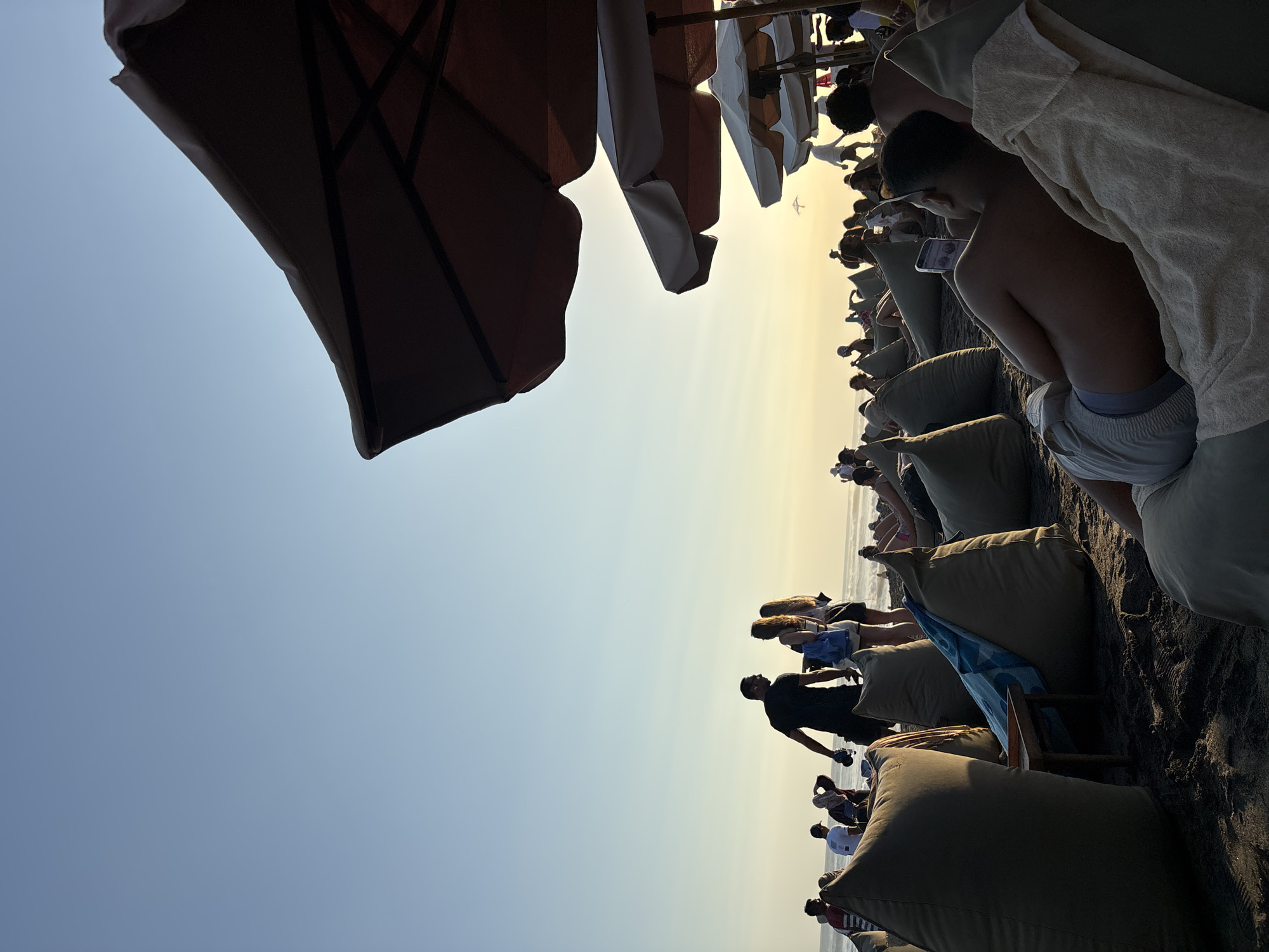 The image captures a crowded beach scene during sunset in North Kuta, Indonesia. The sky displays a gradient from light blue at the zenith to a warm, golden-orange hue near the horizon, indicating the sun's low position. The foreground and midground are dominated by numerous olive-green beanbag chairs placed on dark, wet sand. In the immediate foreground on the right, a shirtless individual lies prone on a beanbag, facing right, looking at a smartphone. A cream-colored towel covers their legs and the beanbag. To their left, a small, low wooden table sits between two beanbag chairs, one of which has a blue and white patterned towel draped over it.
In the midground, various people are scattered across the beach. On the left, a man in a dark t-shirt, holding an object, stands facing two women. Other individuals in silhouette are standing and sitting further to the left. On the right side of the frame, several large beach umbrellas are visible, with the closest two being dark brown and partially obscuring the sky. Further back, lighter-colored umbrellas are seen alongside more people seated on beanbags. In the background, the ocean appears as a dark band at the horizon, with faint wave patterns. Numerous silhouettes of people are visible walking or standing along the distant shoreline. A small, dark, kite-like object is discernible against the bright horizon in the upper-mid-right section of the image.