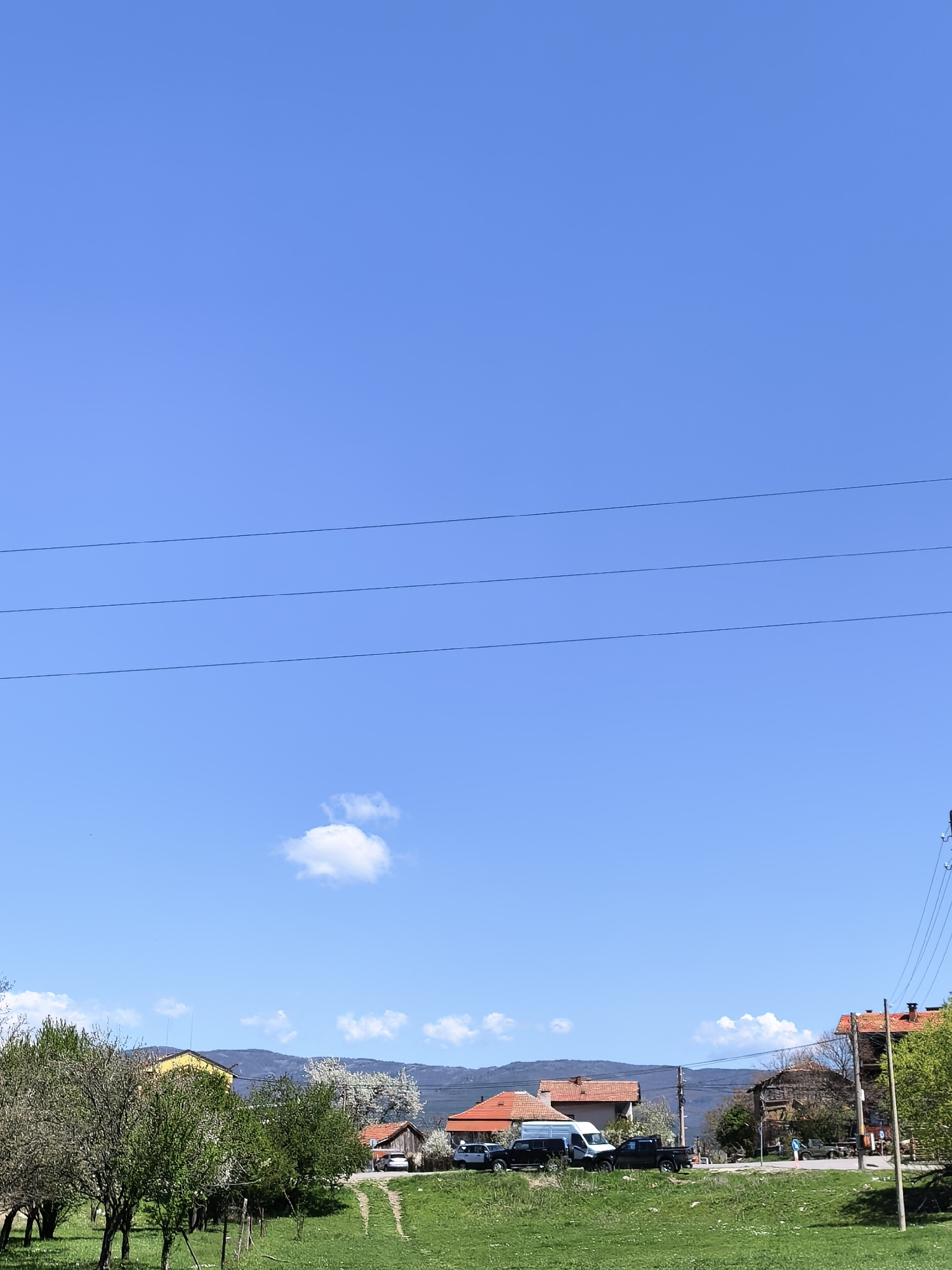 The image depicts a rural or suburban landscape under a clear blue sky with a few wisps of clouds. In the foreground, there's a grassy field with visible tire tracks suggesting vehicle passage. Trees and shrubs line the left edge of the field.
In the midground, a row of houses with red tile roofs is visible, along with parked vehicles including a white van and several trucks. A person is walking along a road. Behind the houses, there are low-lying mountains visible in the distance.
Power lines stretch horizontally across the sky in the upper portion of the image. An electrical pole is on the right edge of the image.