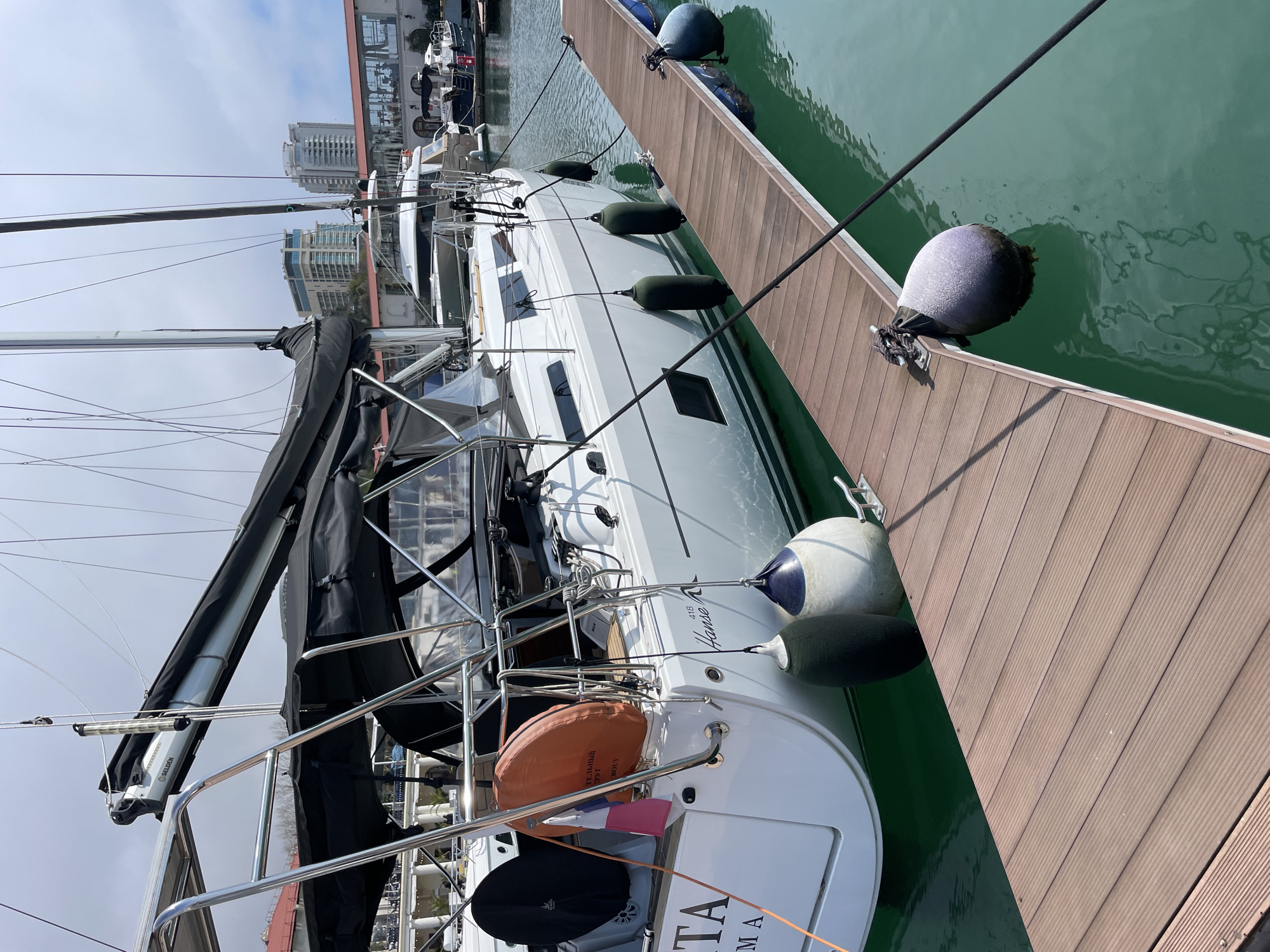 The image shows a white sailboat docked at a wooden pier. The boat has a black sail cover and several fenders hanging along its side to protect it from the pier. The water is a greenish hue. In the background, there are buildings suggesting a city environment, potentially Sochi, Russia. The city is on a harbor. The photograph appears to have been taken on a sunny day.