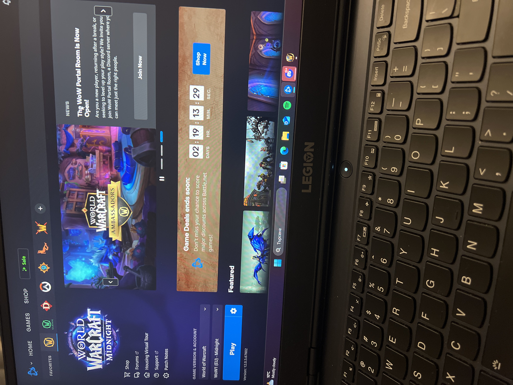 The image shows a black Lenovo laptop displaying the Battle.net application, specifically the World of Warcraft page. The application interface shows the World of Warcraft Midnight game selected, news about the "WoW Portal Room", a "Game Deals" countdown, and a "Featured" section. Icons for various games are visible on the left side of the screen. The keyboard of the laptop is also visible. The computer is running Windows, and a weather icon indicates mostly cloudy conditions. There is no indication in the image that it is taken in Sofia, Bulgaria.