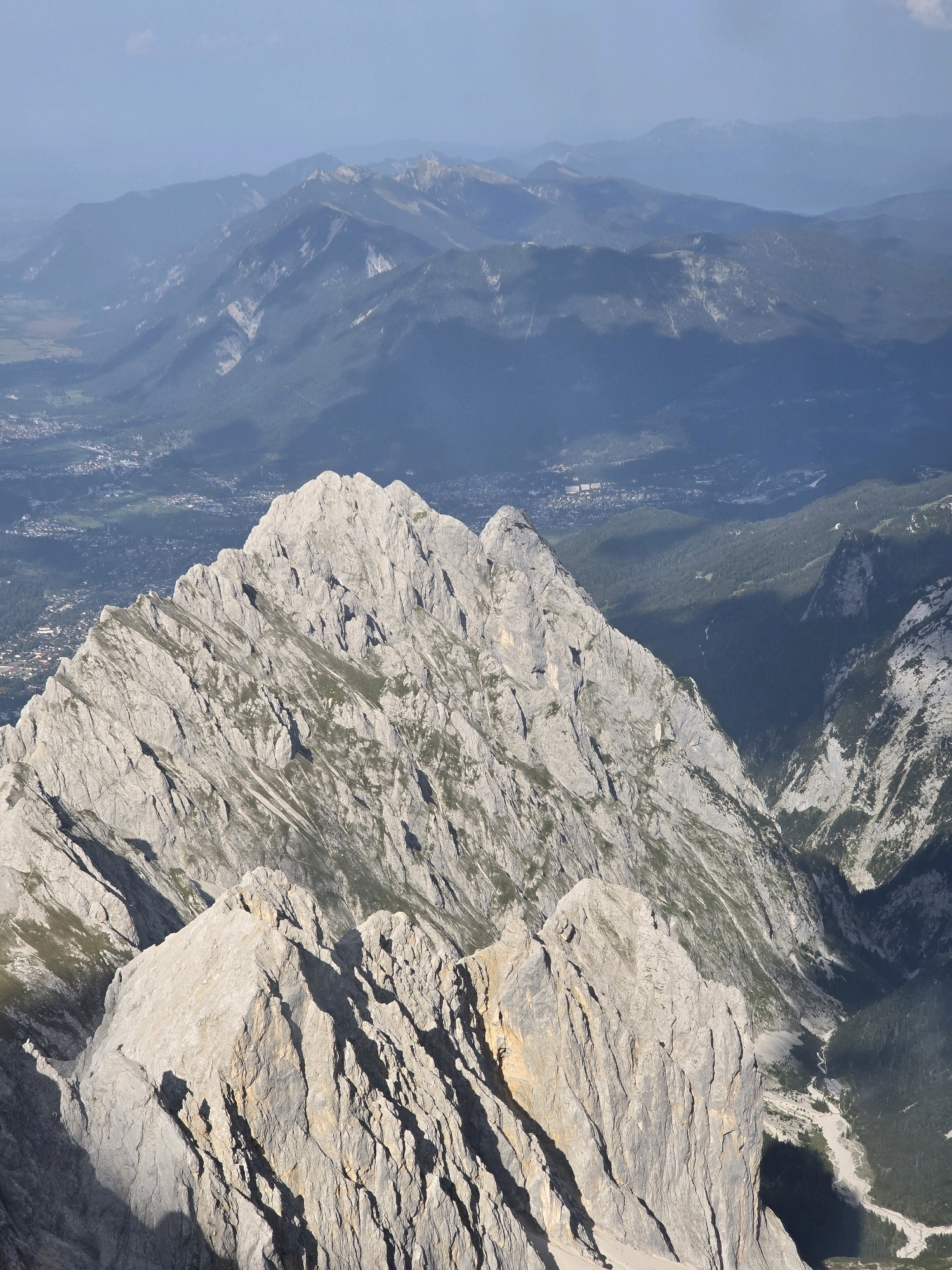The image is a high-angle, wide shot of a mountainous landscape, likely taken from a peak looking down. In the foreground, jagged, light-colored rock formations dominate, with deep shadows cast across their surfaces. Sparse patches of green vegetation are visible clinging to the rock faces.
In the middle ground, a sharper, more defined mountain peak with a similar rocky texture is prominent. Beyond this, a valley opens up, revealing a town or village with buildings and infrastructure. The town is nestled amongst lush green forests that climb the slopes of the surrounding mountains.
In the background, layers of mountains recede into the distance, becoming increasingly hazy and blue due to atmospheric perspective. The sky is a pale blue with some scattered light clouds. The overall scene conveys a sense of vastness and natural grandeur. The presence of a town suggests this is a location near human habitation within a significant mountain range. Based on the provided context, this scene is identified as being near the city of Grainau, Germany, a region known for its alpine scenery.