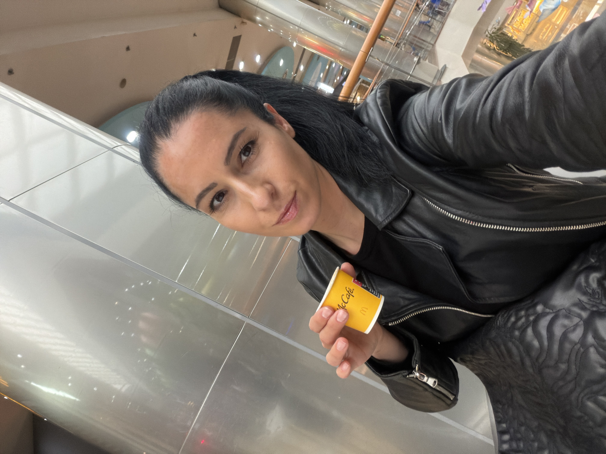 The image captures a woman taking a selfie in an indoor, well-lit modern environment, likely a shopping mall or a transport hub, possibly in Pleven, Bulgaria. She is positioned at an angle, looking directly into the camera with a subtle, confident smile. Her dark hair is pulled back in a ponytail, and she wears a black leather jacket over a dark top. Her nails are short and natural.
In her right hand, she holds a small, yellow paper cup, clearly displaying the "McCafe" brand name and the McDonald's "M" logo in red and white. The background features bright, reflective surfaces, possibly aluminum or glass panels, and hints of overhead lighting fixtures. Reflections of architectural elements and distant lights are visible on these surfaces. The overall impression is one of a casual moment captured in a clean, contemporary public space. The time of day is not discernible from the indoor lighting.