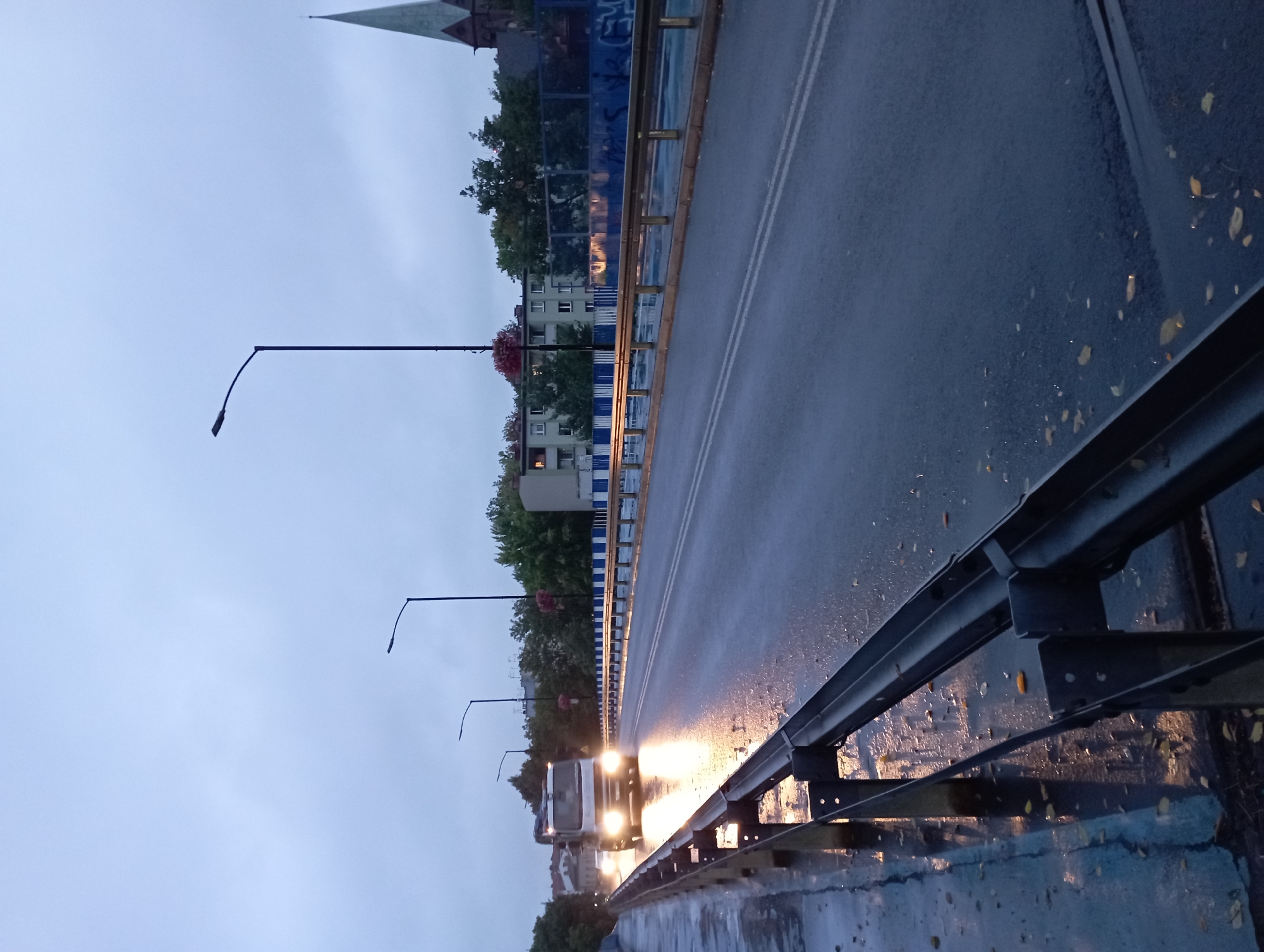 This image depicts a street scene on a bridge in Siemianowice Śląskie, Poland, during what appears to be twilight or early morning. The road surface is wet, reflecting the headlights of an oncoming truck. The truck, a white flatbed with its headlights on, is the primary moving object in the frame, heading towards the viewer on the right side of the road. The bridge has metal railings on both sides, with a blue and white striped barrier further along the right side. Lampposts are visible along the bridge, and in the background, there are buildings and trees. A church steeple with a dark cross is visible on the far right of the image, characteristic of the architecture in Siemianowice Śląskie. The sky is overcast and a pale gray. Scattered leaves are visible on the wet road surface and the pedestrian walkway. The overall atmosphere is somber and damp, suggesting recent rain.