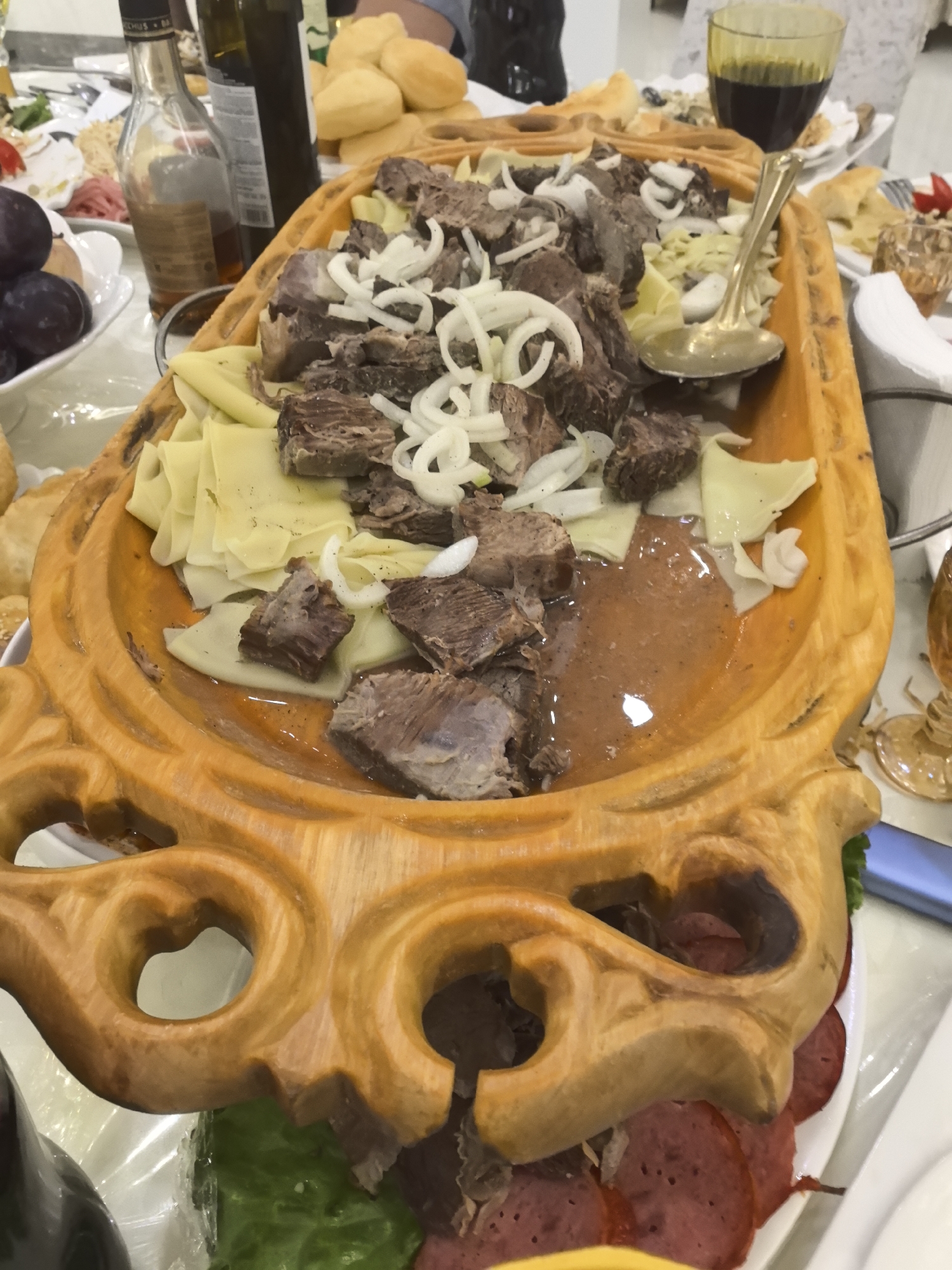 A close-up, overhead shot shows a large, intricately carved wooden platter holding a dish of meat, noodles, and onions, with a spoon resting on top. The platter is ornately decorated with carved swirls and heart shapes. Beneath the platter, a white plate holds sliced salami. The background reveals other dishes, bottles, and a glass of dark liquid, suggesting a meal setting. The overall scene is well-lit, with the food being the central focus.