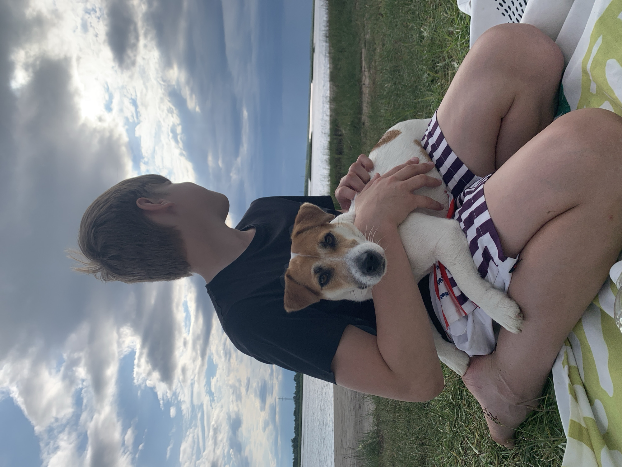 A young person, viewed from the chest up, is sitting outdoors on a grassy area with a beach and body of water in the background. The person is wearing a black t-shirt and striped shorts. They are holding a small, white and brown dog in their arms. The dog is looking directly at the camera. The sky is filled with clouds. This image was taken in Severodvinsk, Russia.
