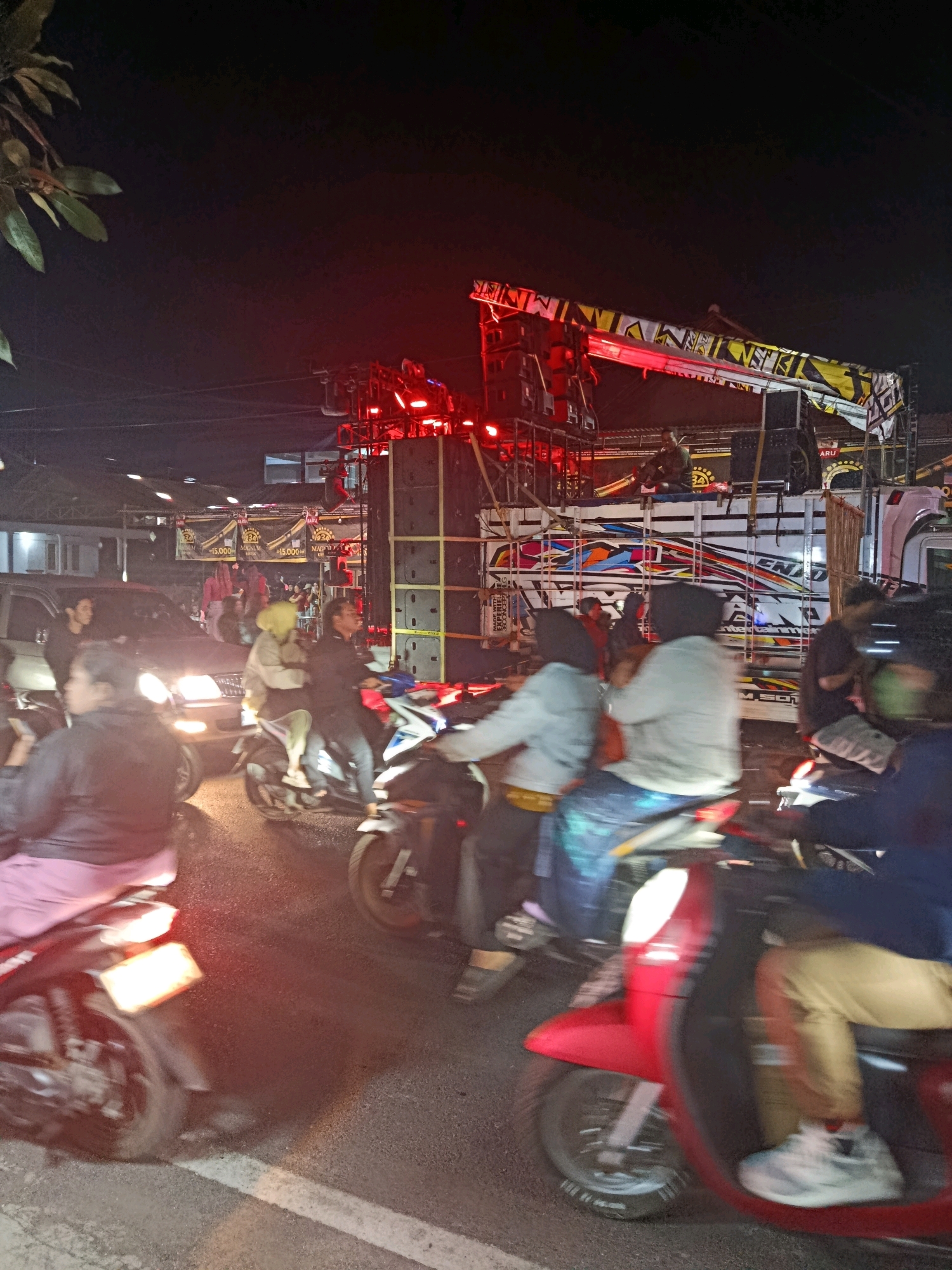 An outdoor nighttime scene in Turen, Indonesia, features a loud sound system mounted on a decorated truck, with several people on motorcycles in the foreground. The truck is adorned with colorful banners and speakers, and a person is seated on top of it. The road is asphalt, and there are buildings and lights visible in the background, suggesting an urban setting. People on motorcycles are either stopped or moving slowly, some with passengers. The overall atmosphere appears to be festive or event-like, with the prominent sound system indicating a possible music event or celebration.